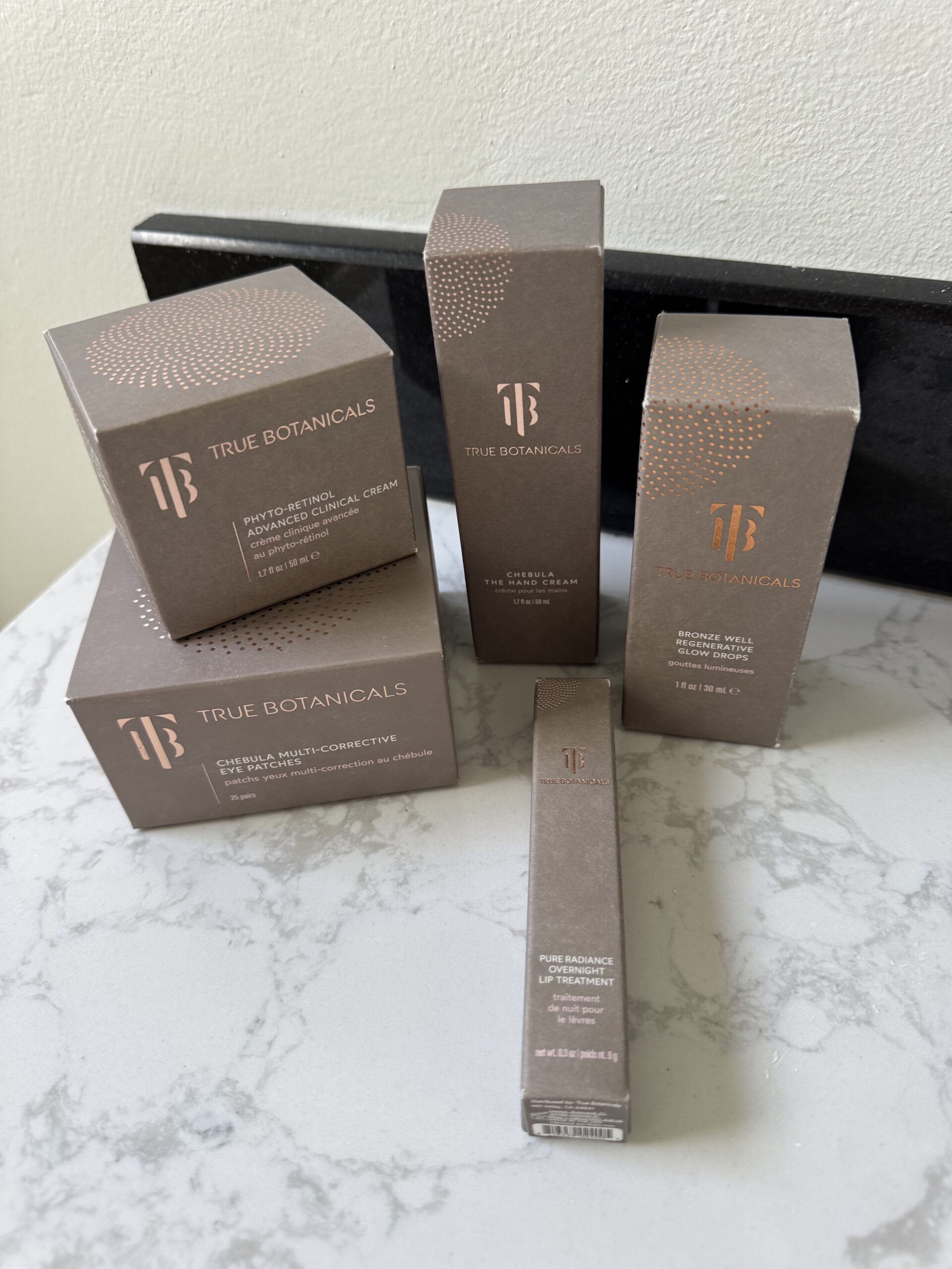 Five True Botanicals skincare product boxes are arranged on a marble surface against a light-colored wall.