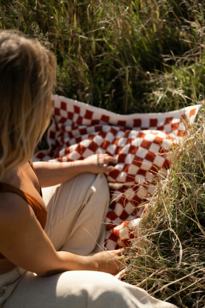 A person sits on dry grass next to a red and white checkered blanket, wearing a brown top and light pants, partially turned away from the camera.