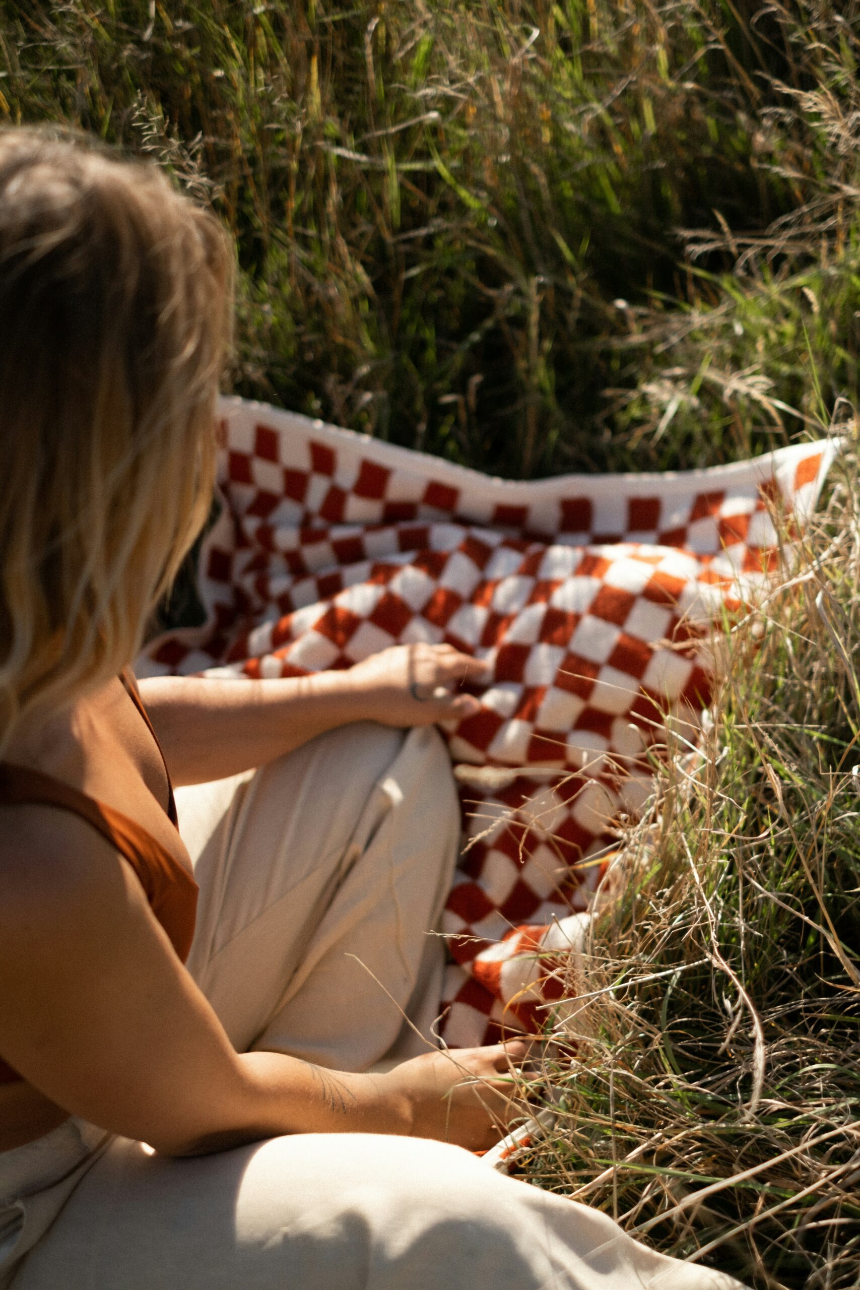 A person sits on dry grass next to a red and white checkered blanket, wearing a brown top and light pants, partially turned away from the camera.