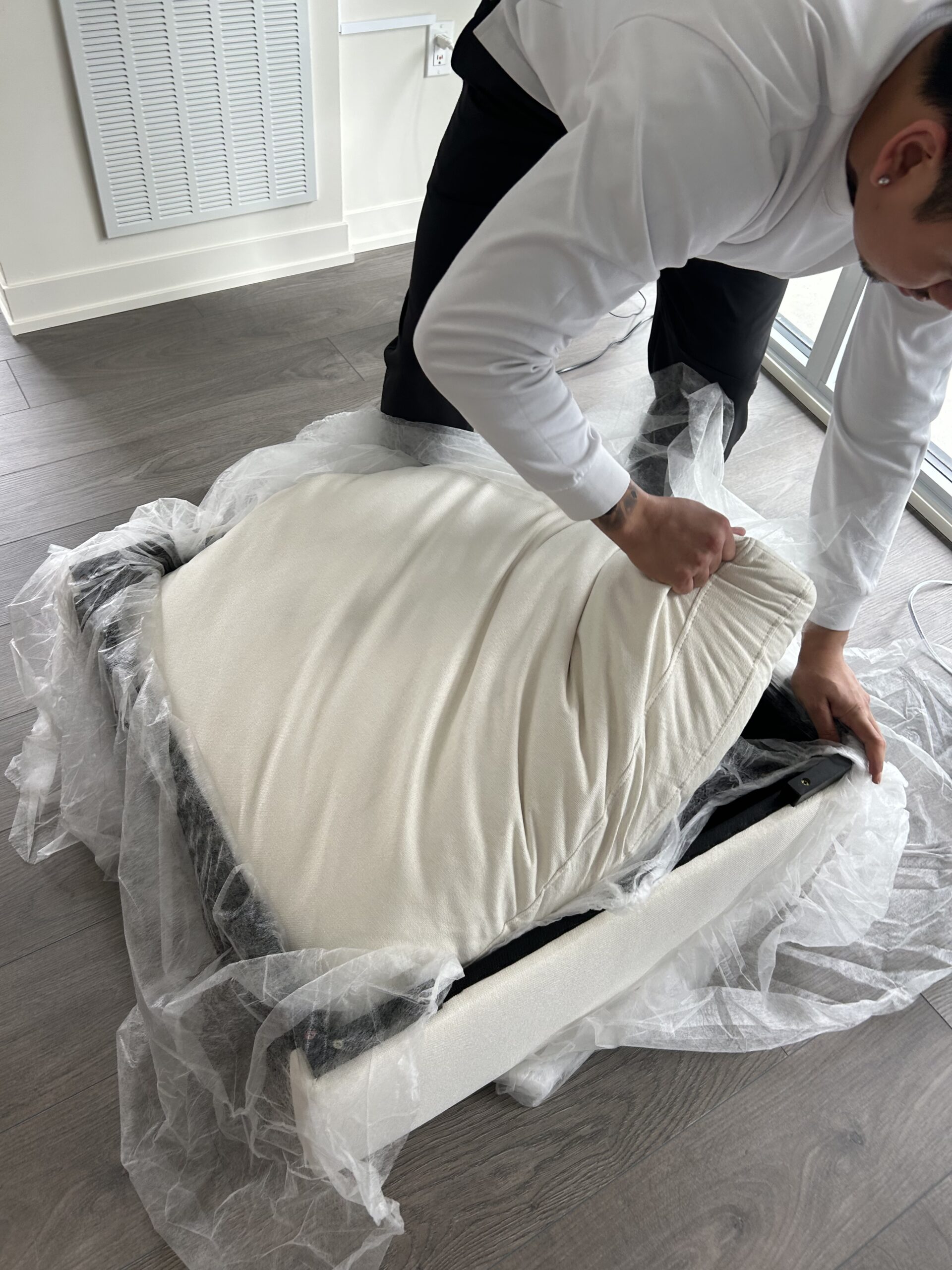 A person in a white shirt and black pants unwraps a cushioned chair seat from protective plastic packaging in a room with wood flooring.