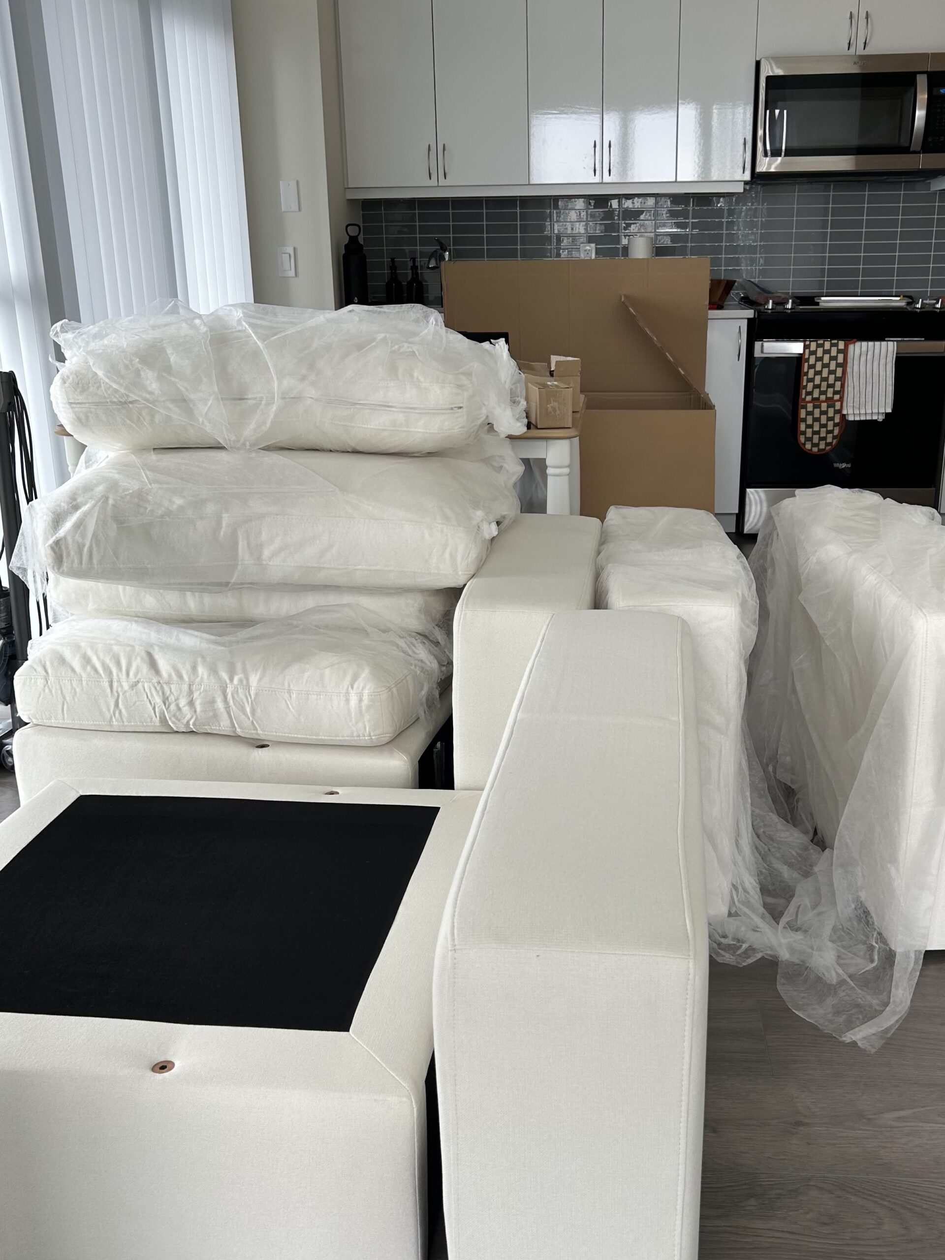 Couch cushions and sections are stacked and wrapped in plastic in a kitchen area, with an open cardboard box and various kitchen appliances visible in the background.