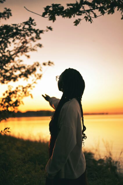 A person with long braids and glasses stands by a lake at sunset, reaching out toward the horizon, with trees framing the scene.