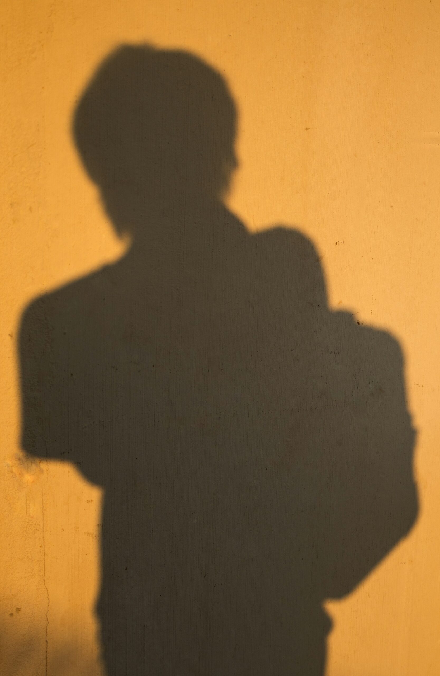 A person's shadow is cast on a yellow wall, with the outline showing them carrying a bag on one shoulder.