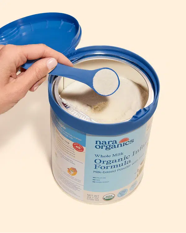 A hand scoops powdered infant formula from an open container of Nara Organics Whole Milk Organic Infant Formula with a blue measuring spoon.