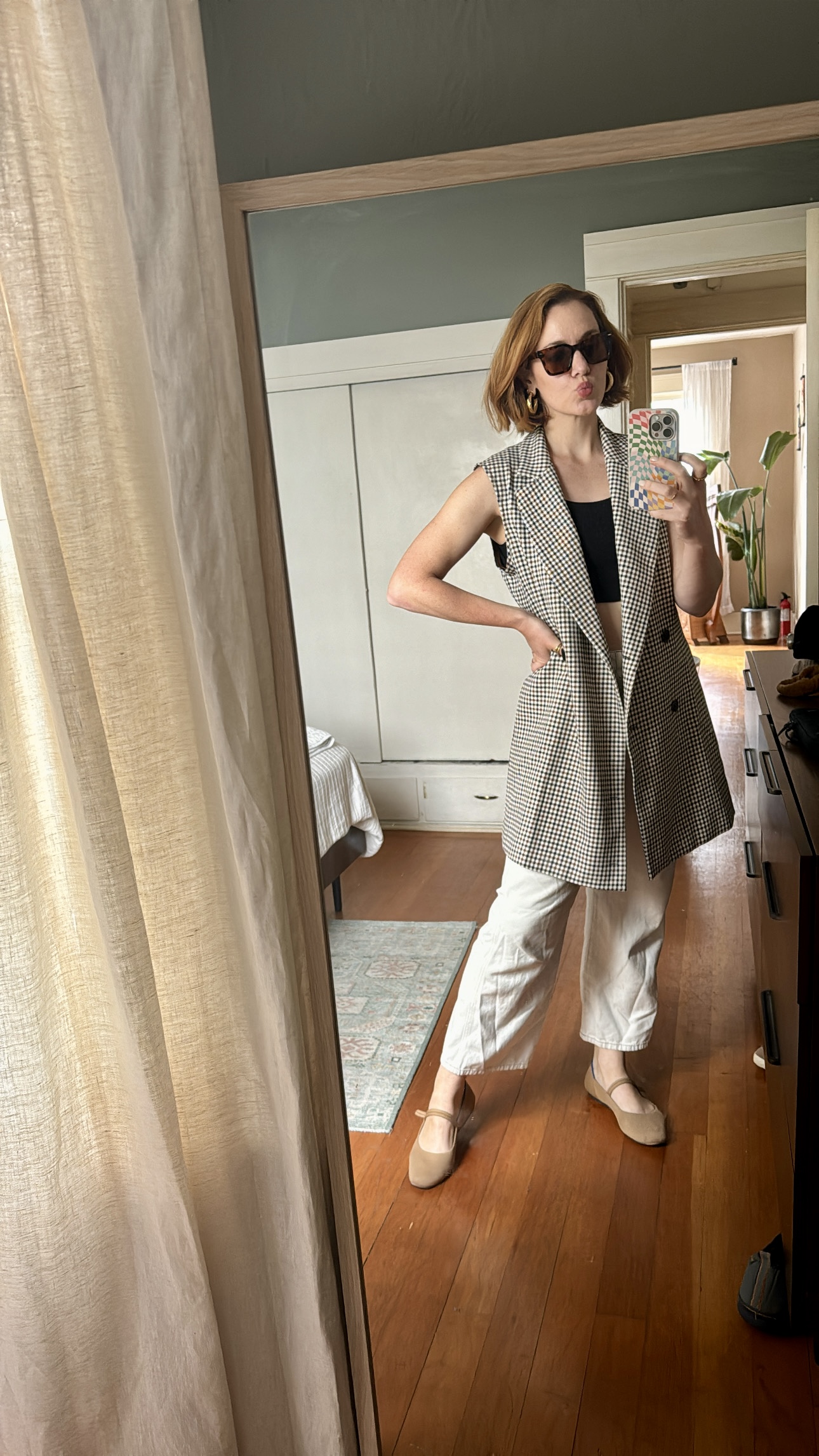 Person stands in front of a mirror taking a selfie, wearing sunglasses, a sleeveless checkered blazer, white pants, and beige shoes in a bedroom with wooden floors.