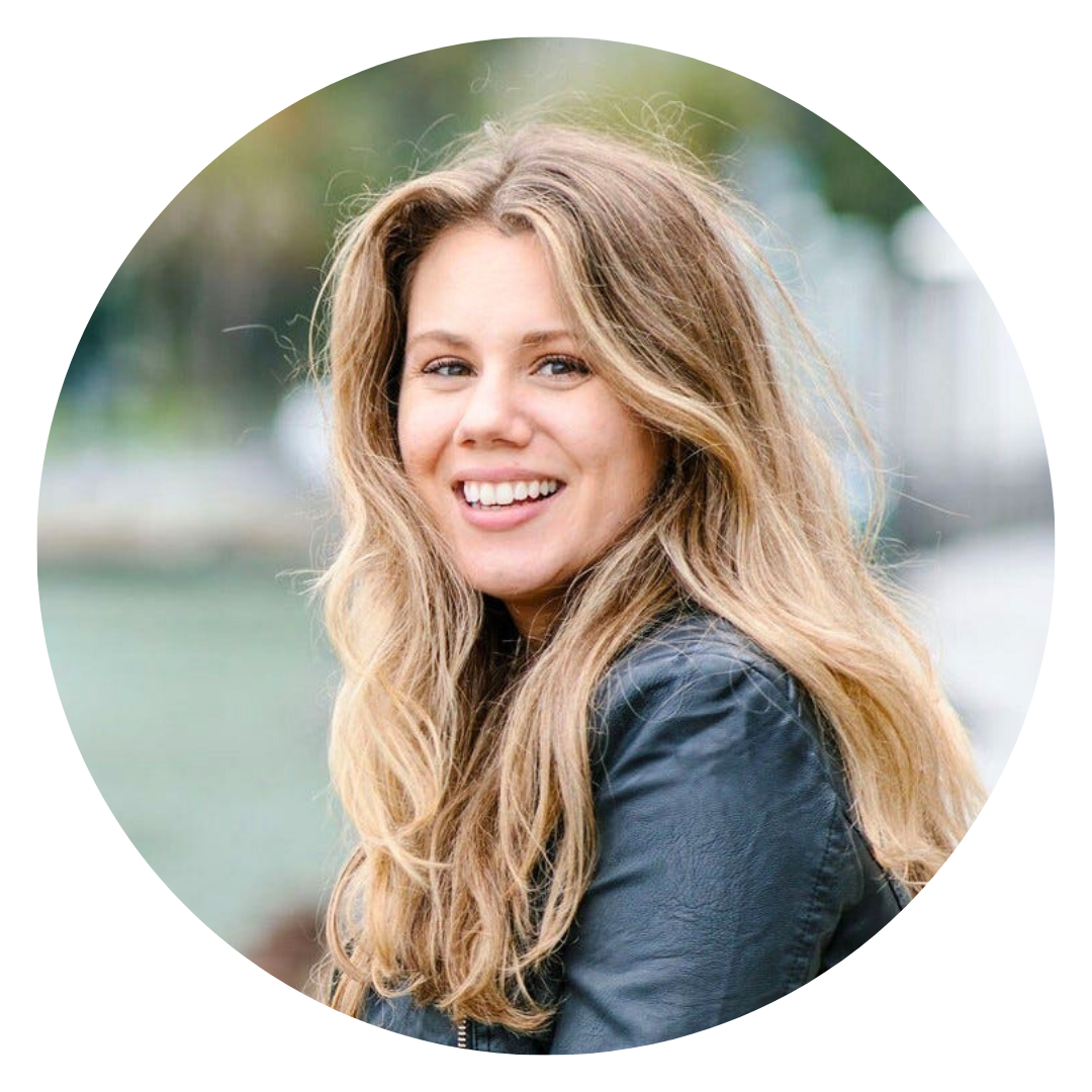 Smiling woman with long blonde hair wearing a black jacket, outdoors with blurred greenery in the background.