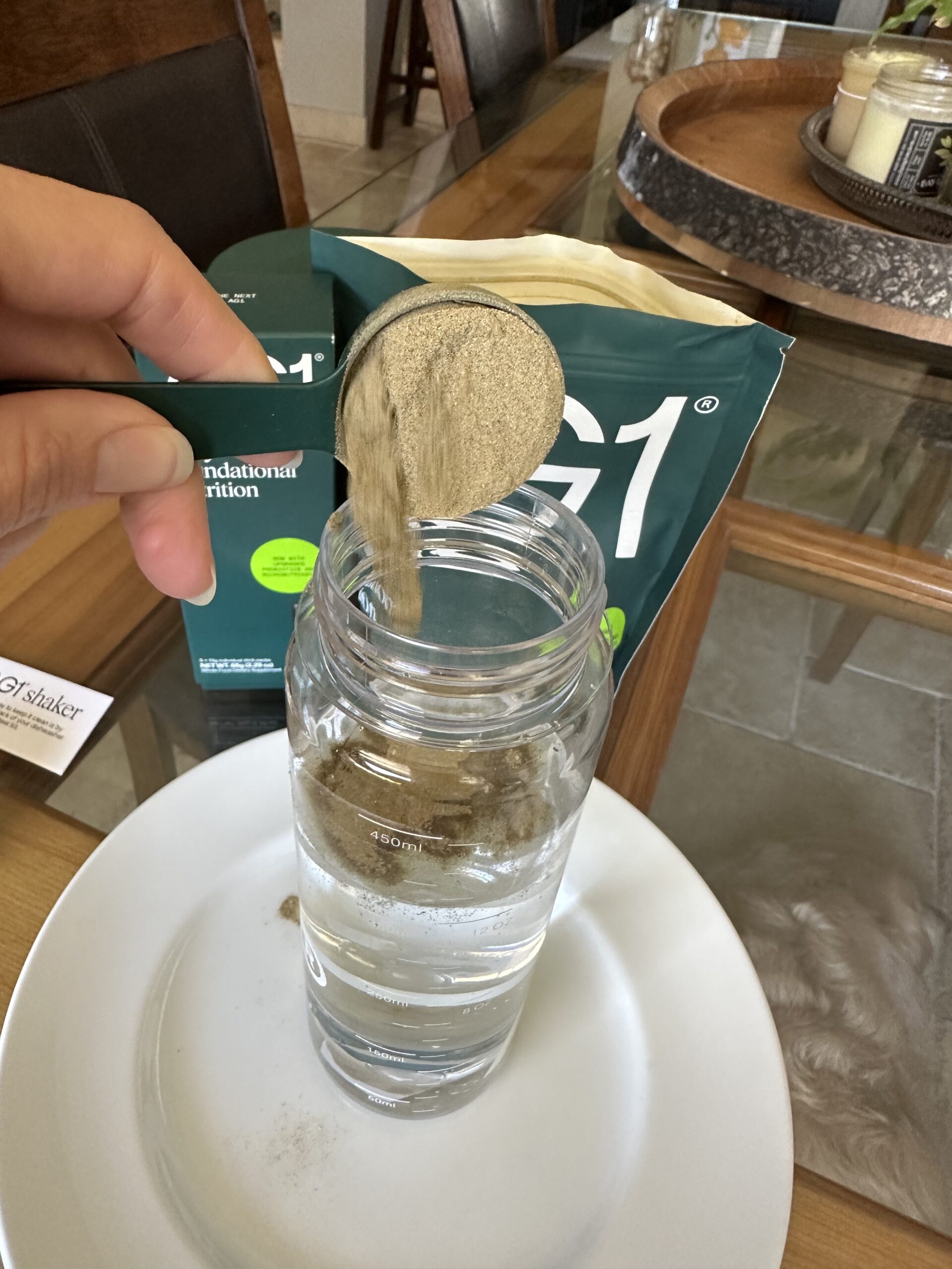 A hand pours powdered supplement from a scoop into a glass of water, with an AG1 nutrition package in the background on a table.