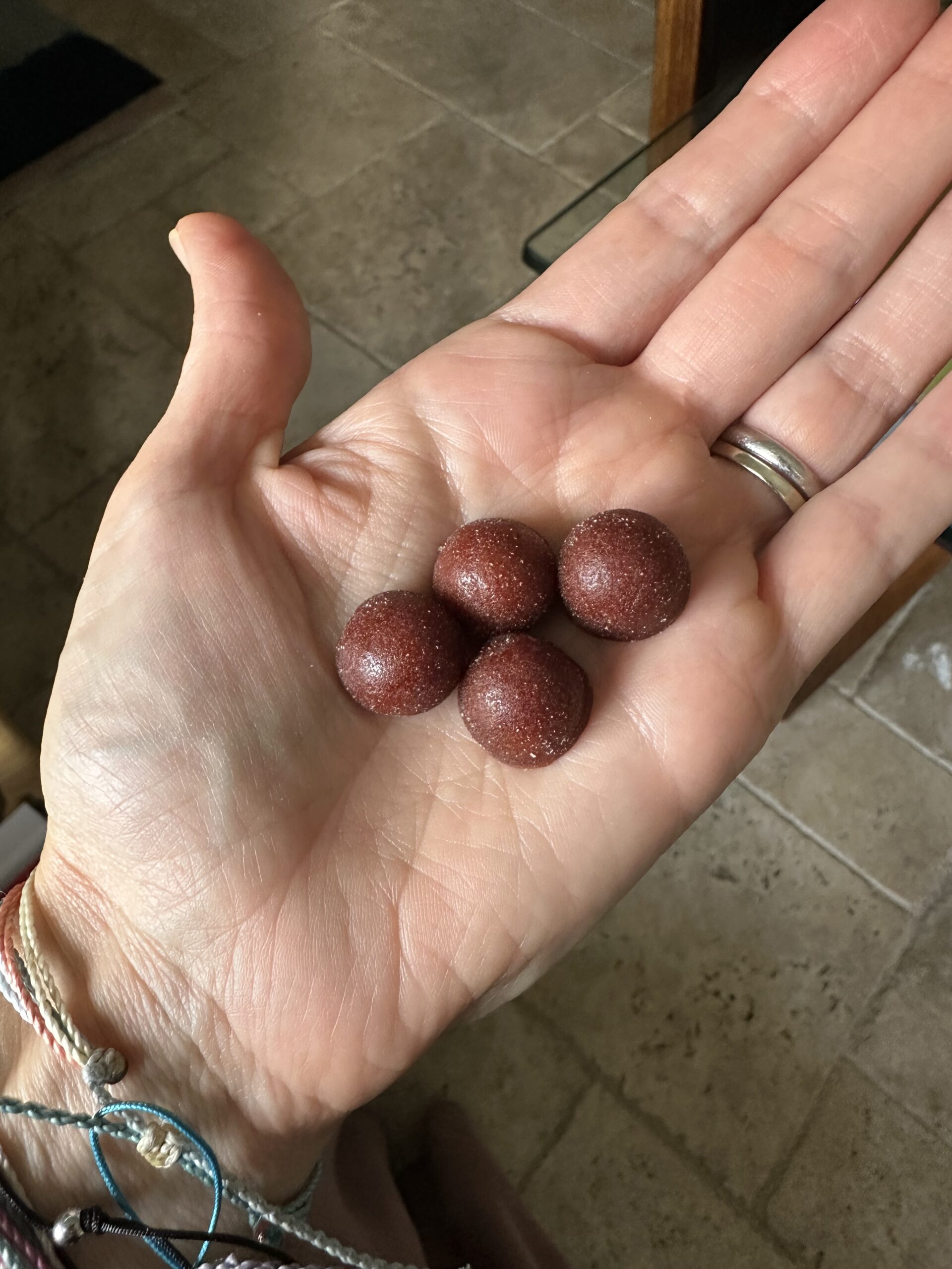A hand holding four round, reddish-brown, sugar-coated candies or gummies over a tiled floor.
