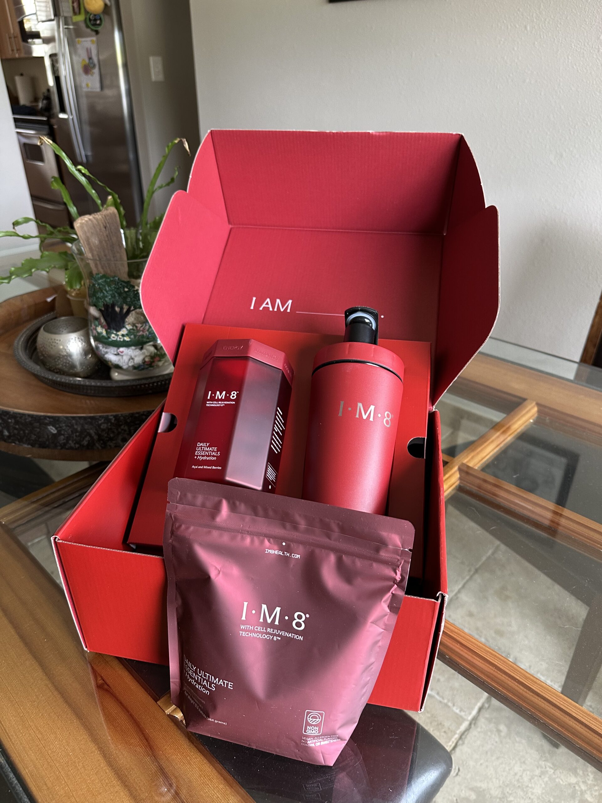 A red box labeled "I AM 8" contains a bottle, a canister, and a pouch of nutritional supplement products on a glass table next to a potted plant.