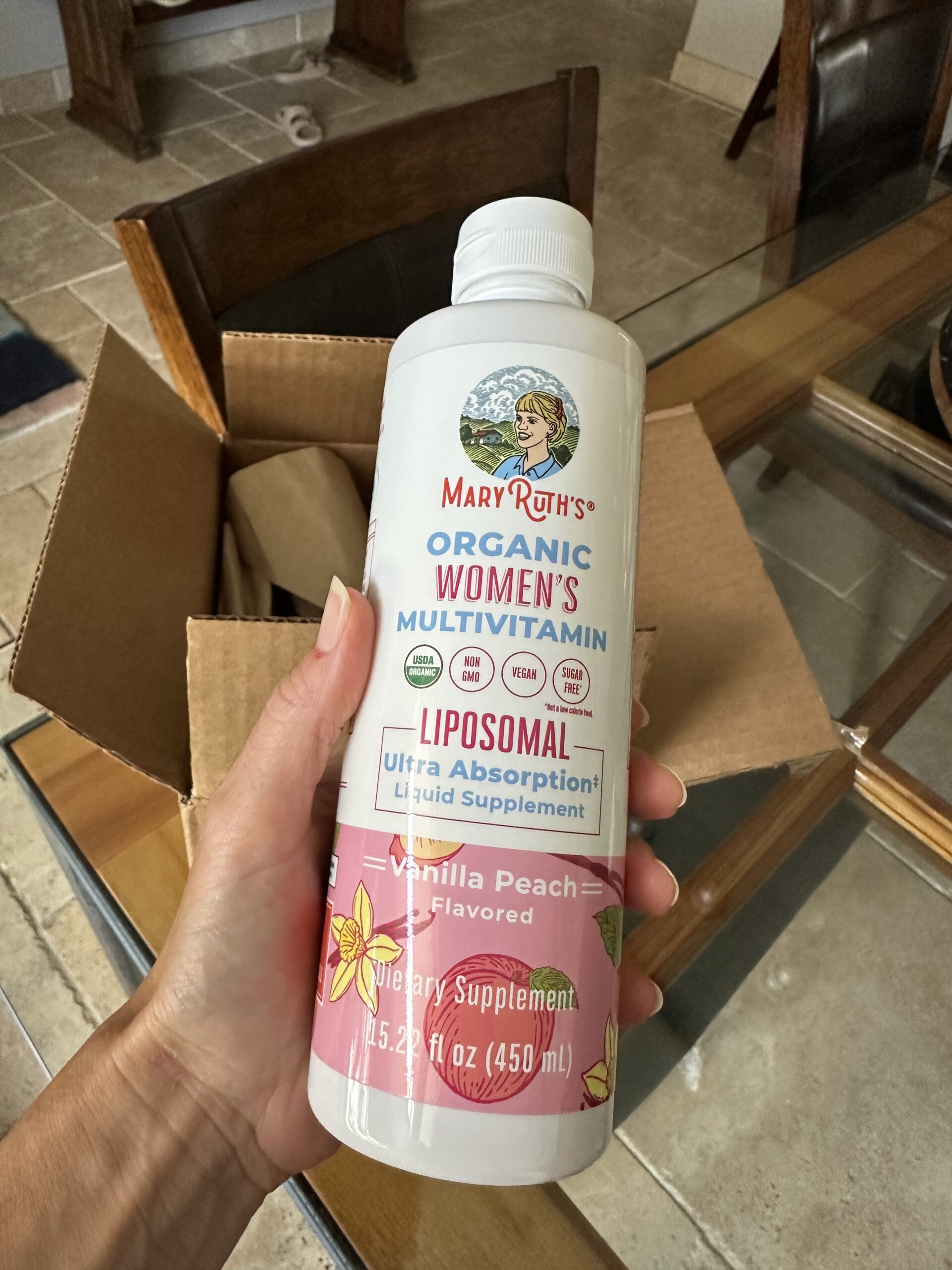 A hand holds a bottle of MaryRuth's Organic Women's Multivitamin Liposomal in vanilla peach flavor, with a cardboard box and kitchen table in the background.