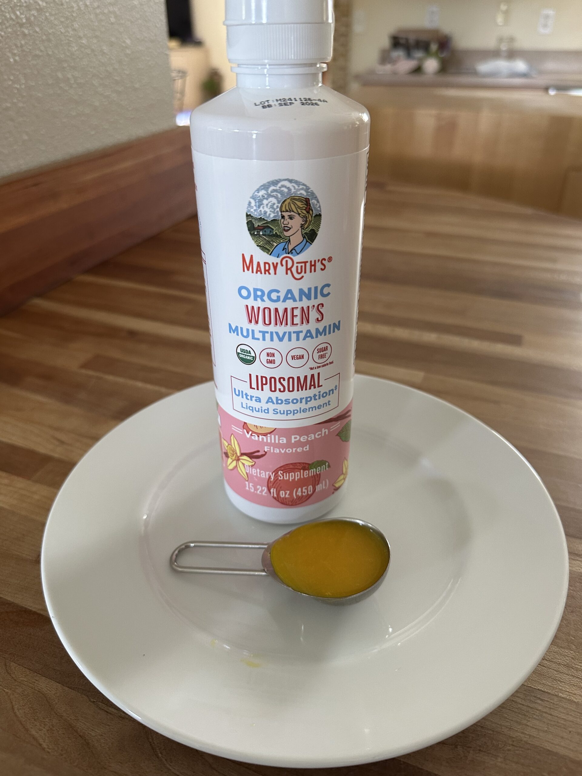 A bottle of MaryRuth's Organic Women's Multivitamin stands on a plate, with a metal tablespoon filled with the liquid vitamin in front.