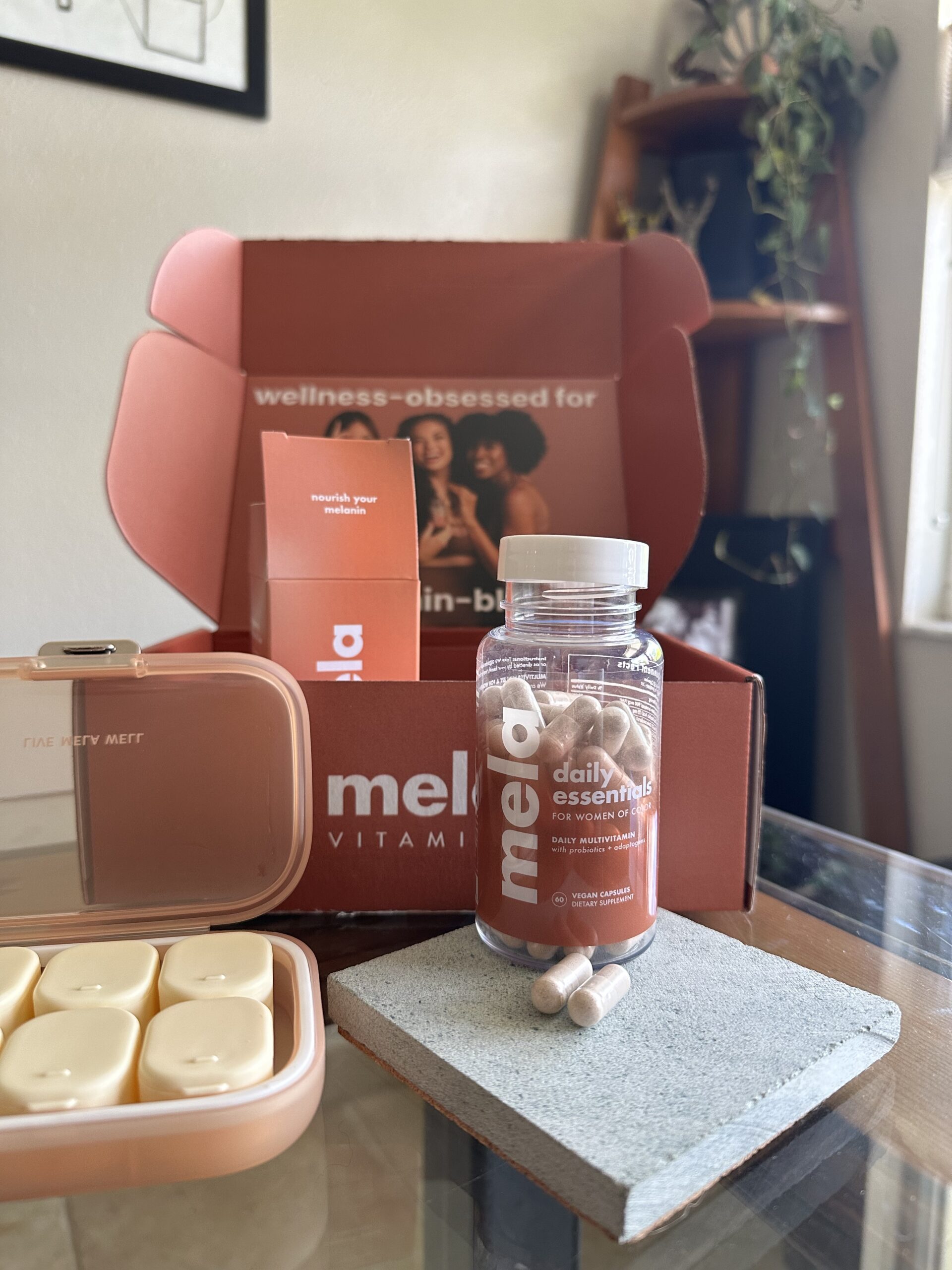 A bottle of daily essential vitamins and a pill case are displayed on a table in front of an open wellness subscription box.