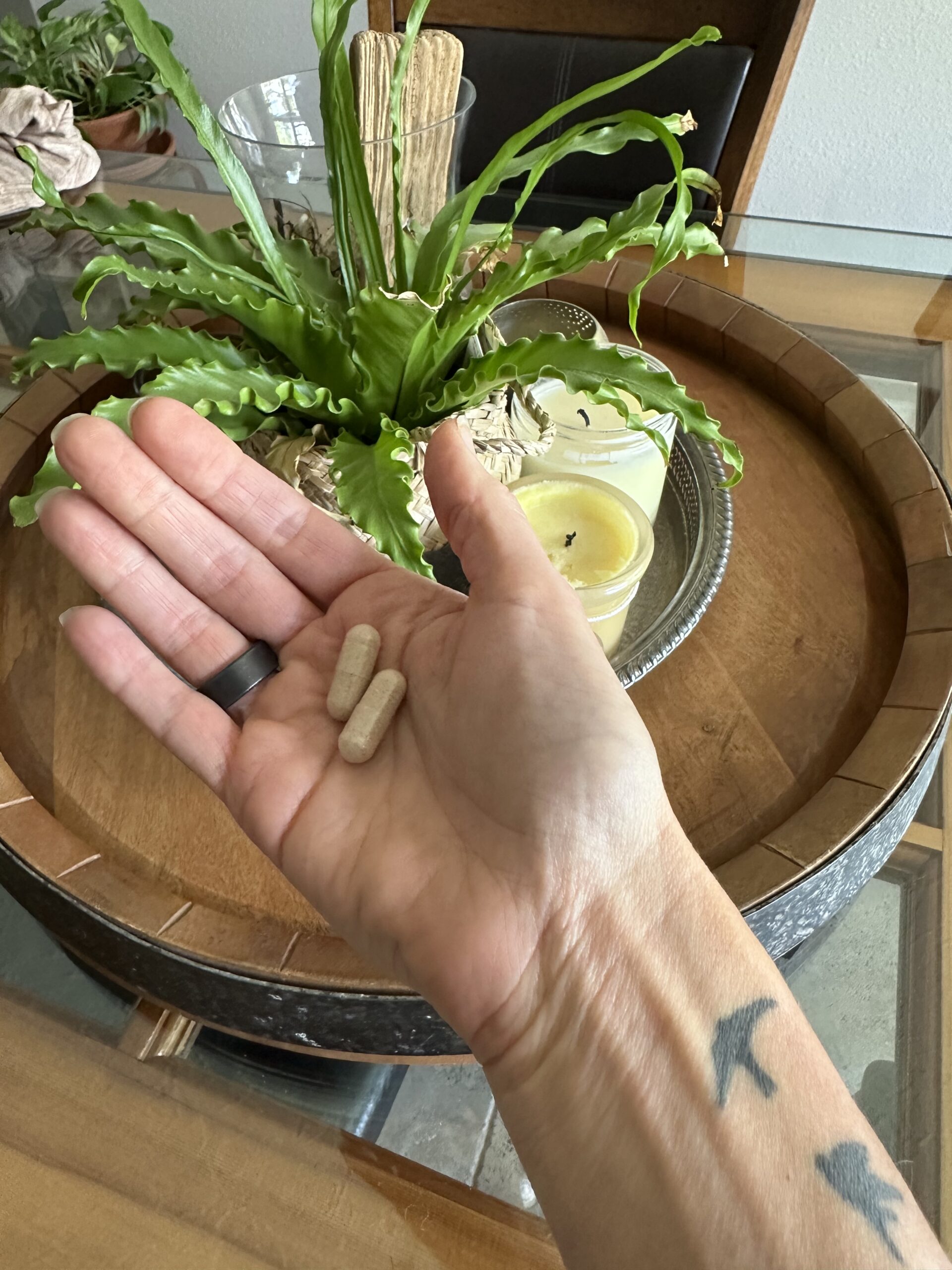 A hand holding two beige capsules above a wooden table with a round tray, candles, and a green potted plant.