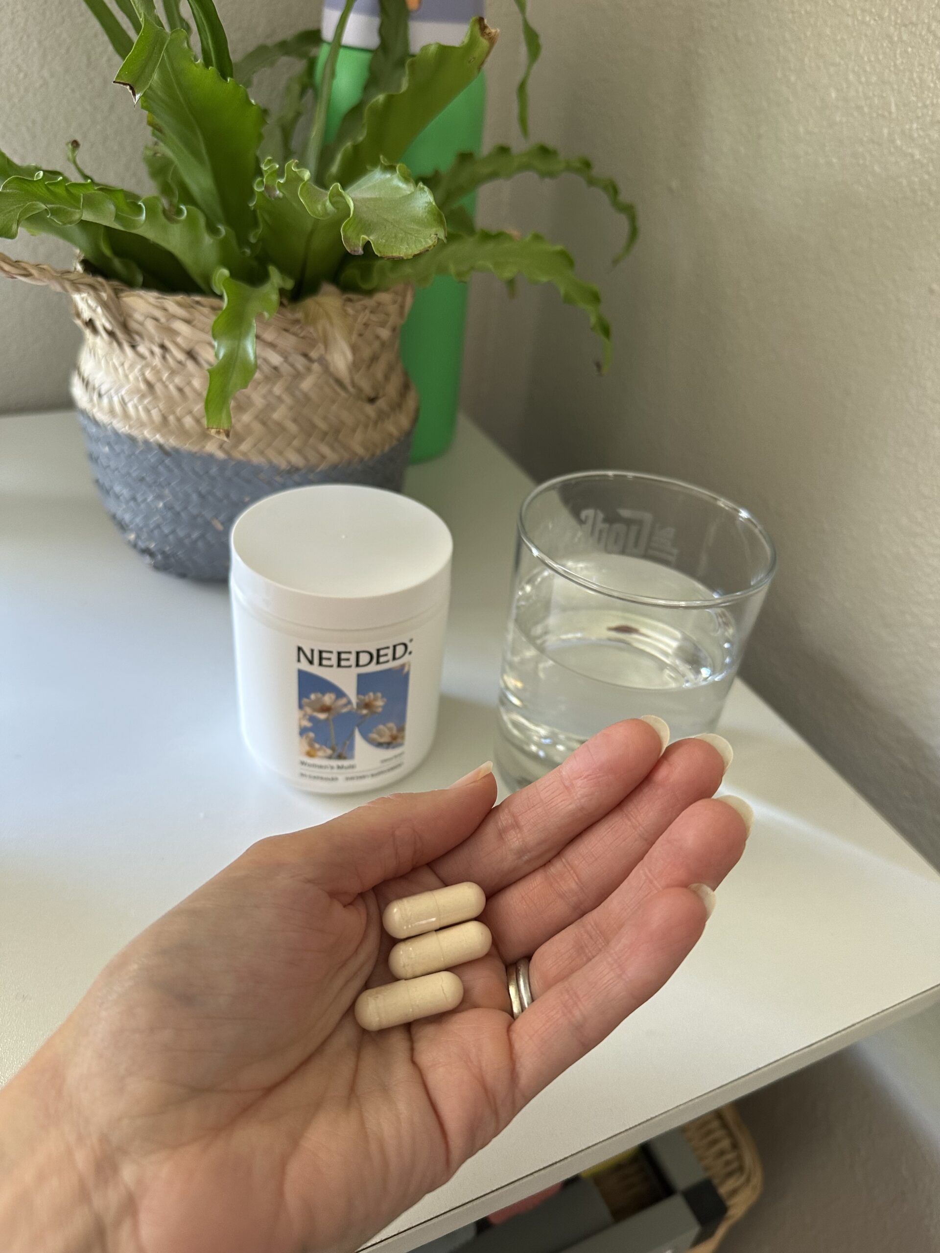 A hand holds three beige capsules above a white surface with a supplement container, a glass of water, and a potted plant in the background.
