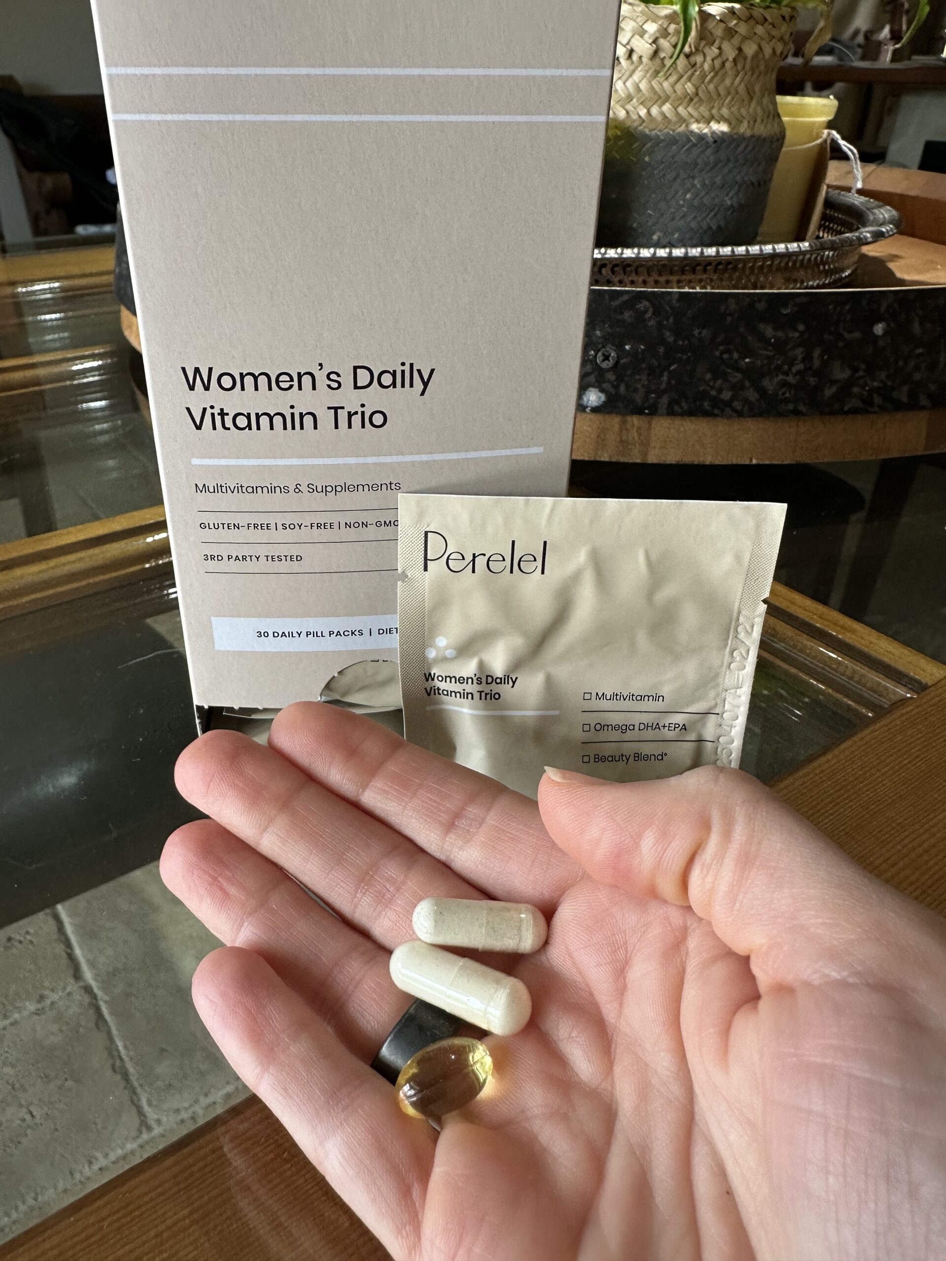 A hand holds four supplement pills in front of a Perel Women's Daily Vitamin Trio box and a packet labeled with the same product name.