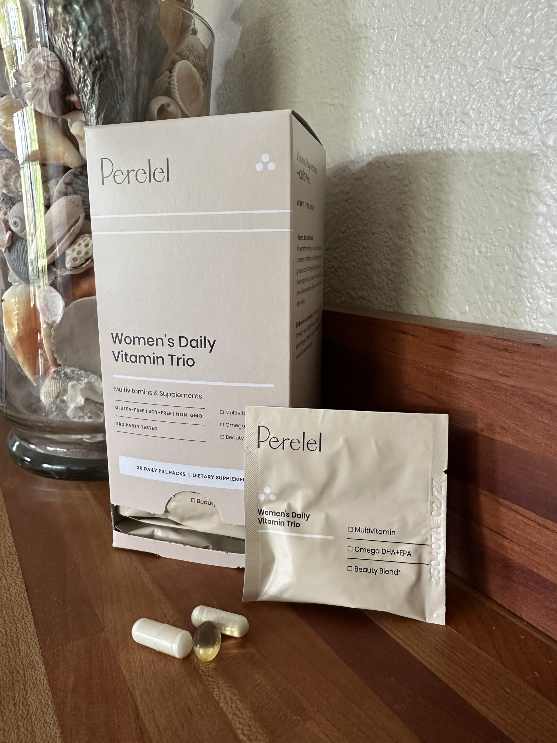 Box and packet of Perelel Women's Daily Vitamin Trio on a wooden surface, with three supplement capsules and a glass container of seashells in the background.