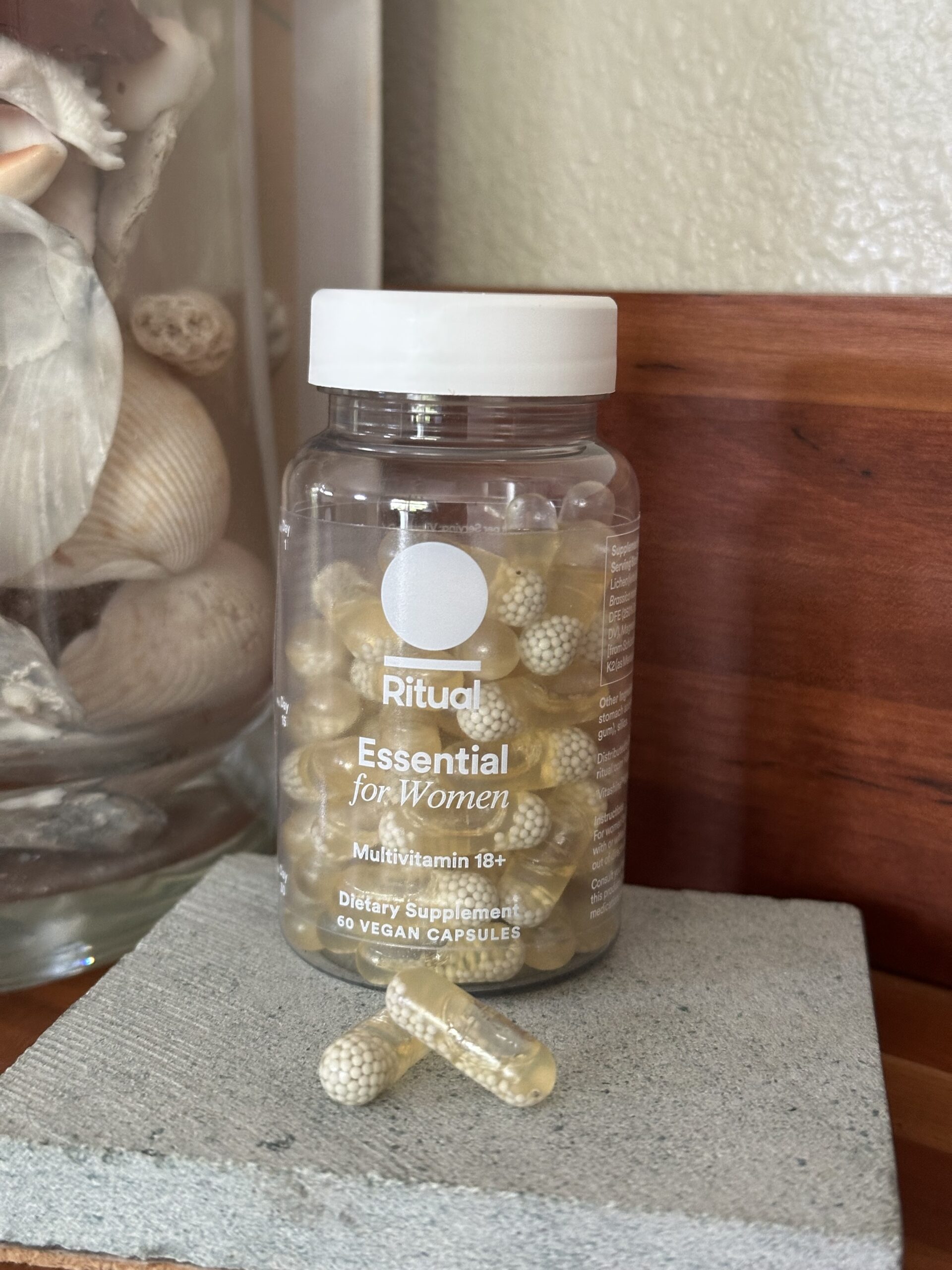 A bottle of Ritual Essential for Women multivitamin capsules, labeled as vegan and for adults 18+, sits on a small gray platform next to a glass container filled with seashells.