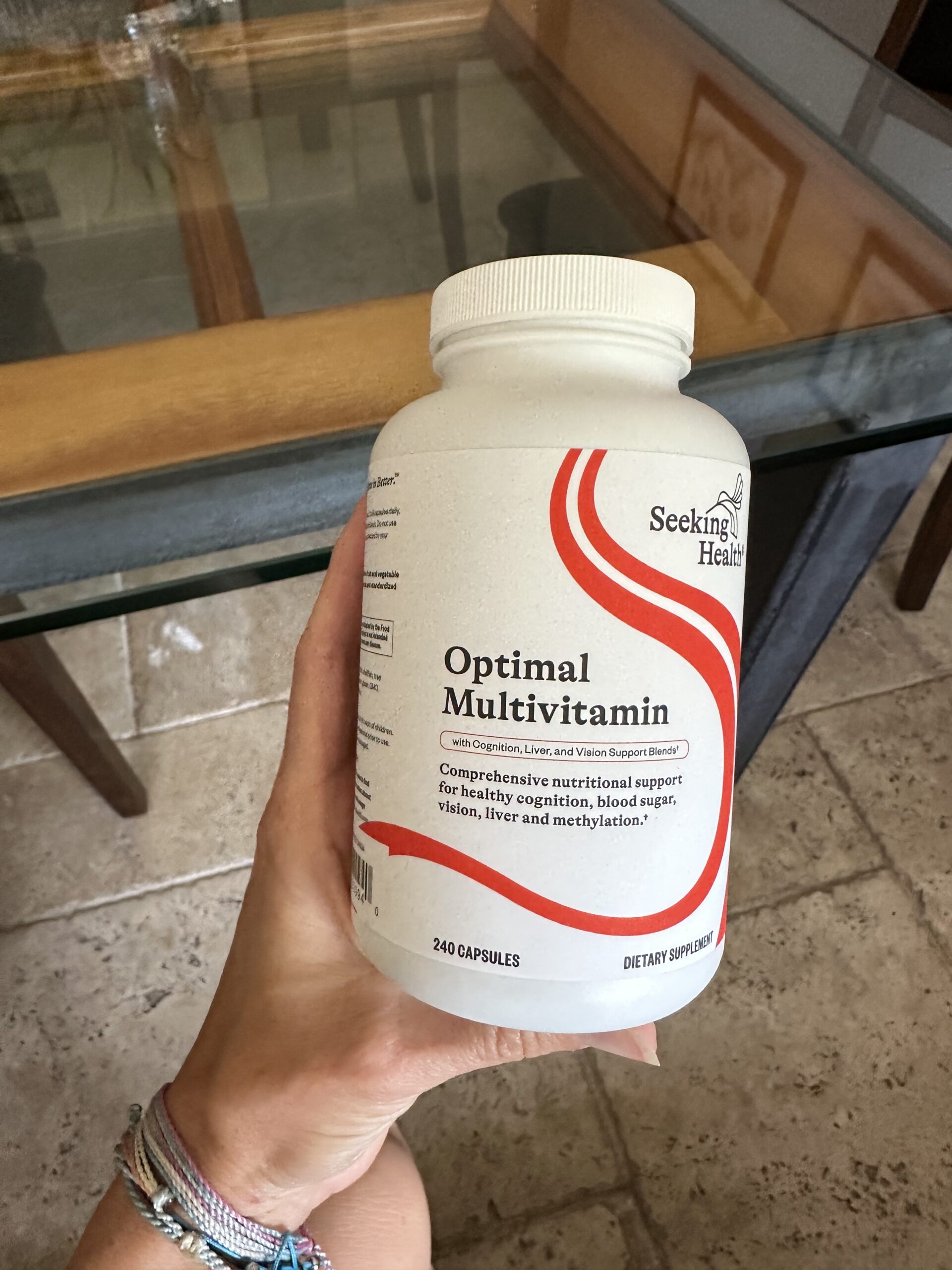 A hand holding a bottle of Seeking Health Optimal Multivitamin containing 240 capsules, with text highlighting support for energy, cognition, blood sugar, vision, liver, and methylation.