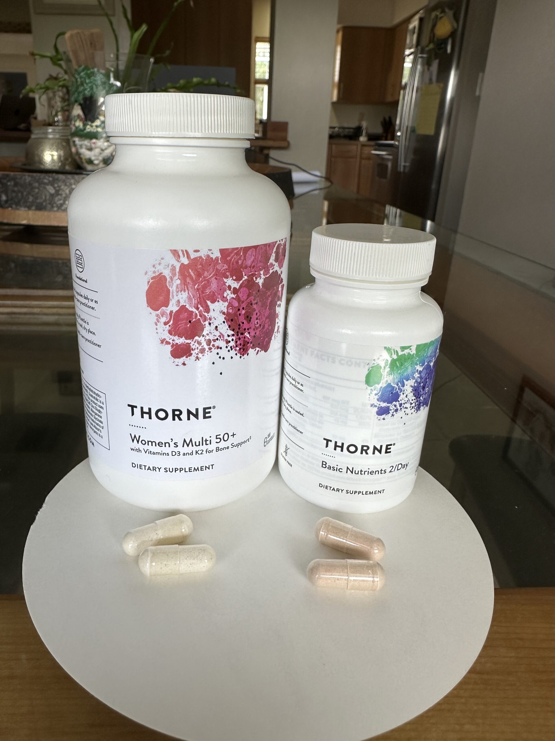 Two bottles of Thorne dietary supplements, Women's Multi 50+ and Basic Nutrients 2/Day, are placed on a table with five assorted capsules in front of them.