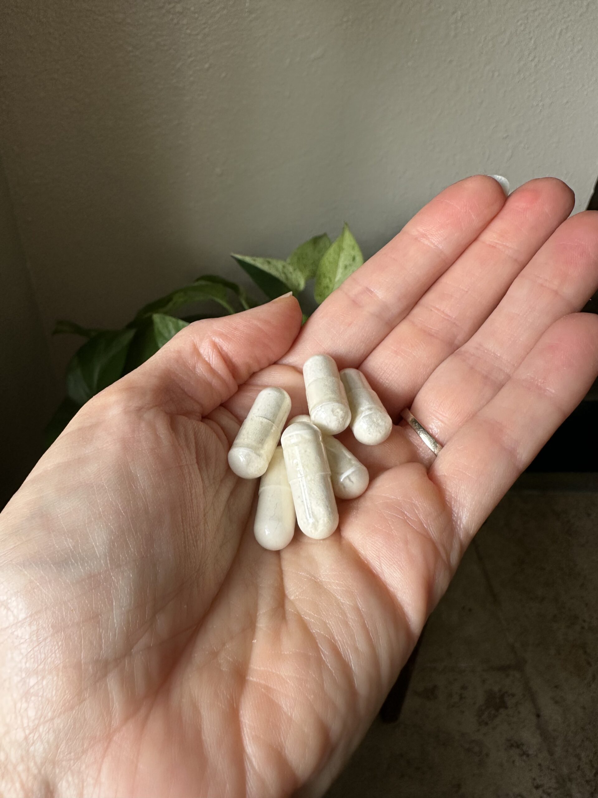 A hand holding six white capsules, with a plant and beige wall in the background.