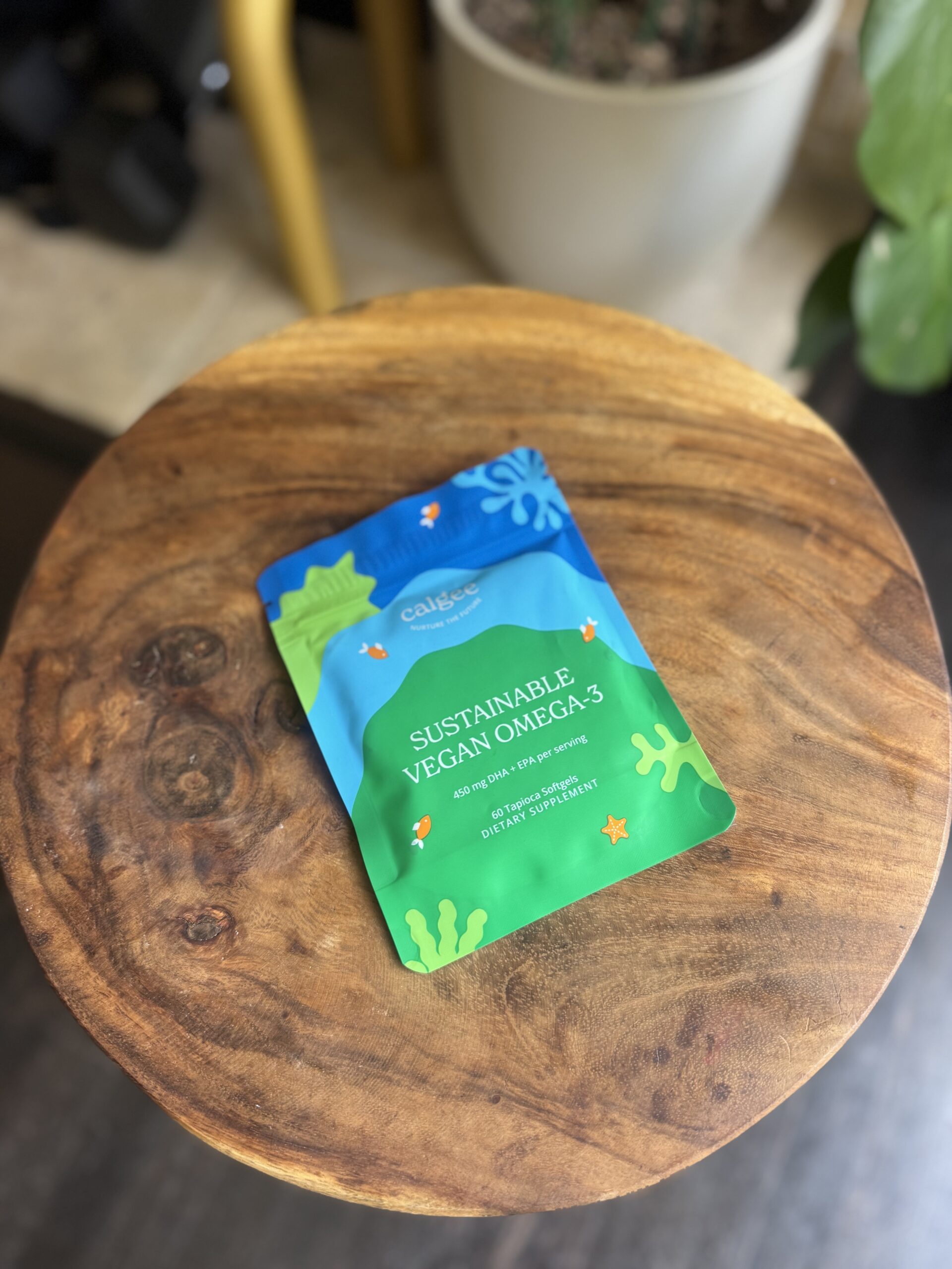 A packet labeled "Sustainable Vegan Omega-3" sits on a round wooden table, with plants visible in the background.