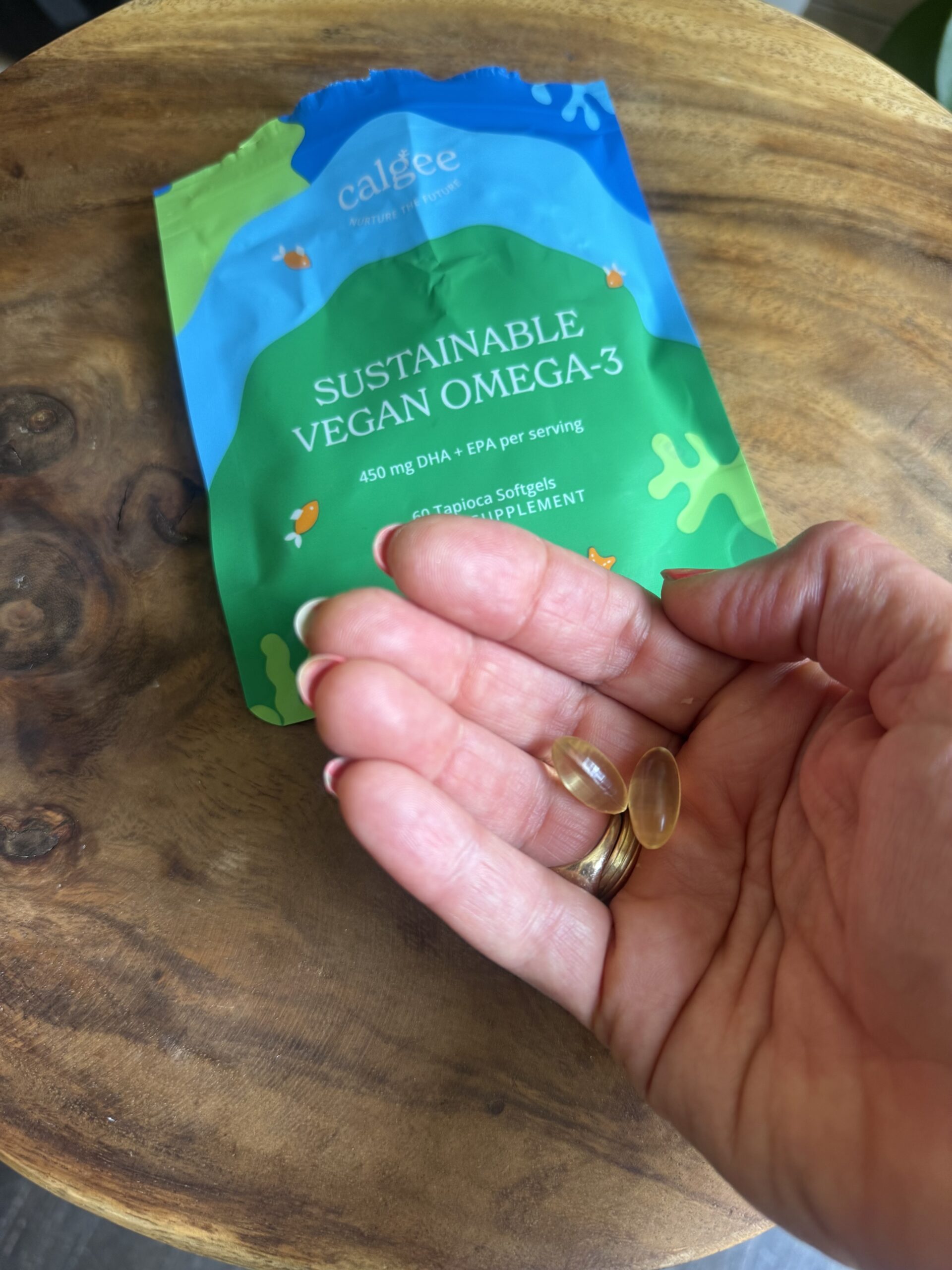 A hand holding two softgel capsules of omega-3 supplement with a "Sustainable Vegan Omega-3" package on a wooden surface in the background.