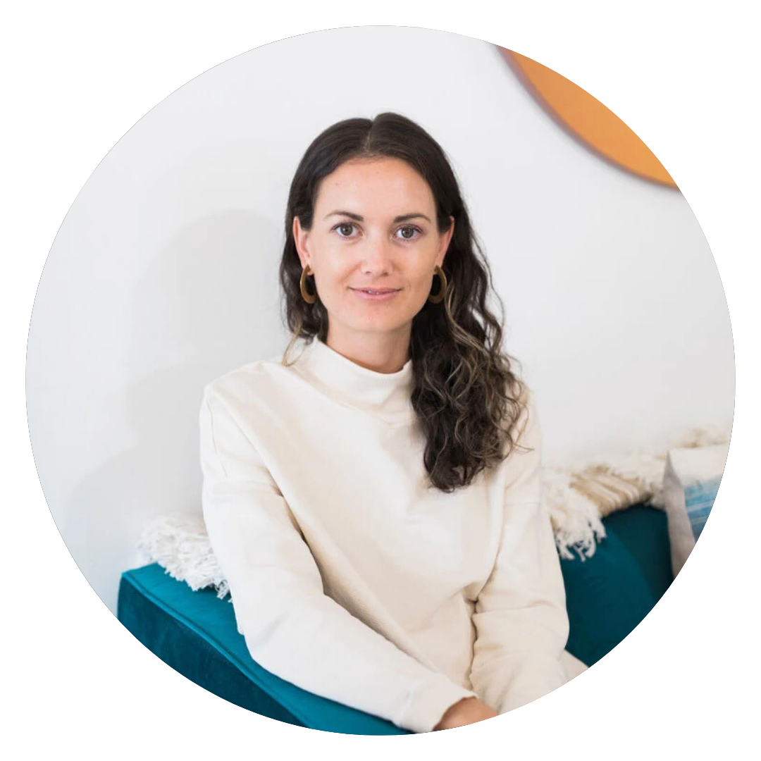 A woman with long dark hair, wearing a cream sweater and gold hoop earrings, sits on a teal couch with a white textured pillow behind her.