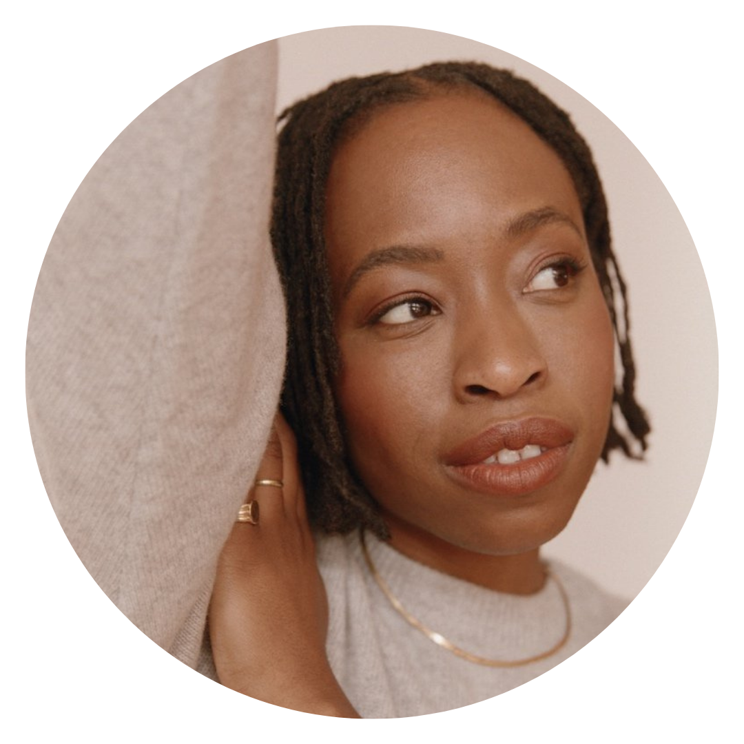 A person with short, twisted hair and gold jewelry looks off to the side with a neutral expression, wearing a light gray top in soft lighting.