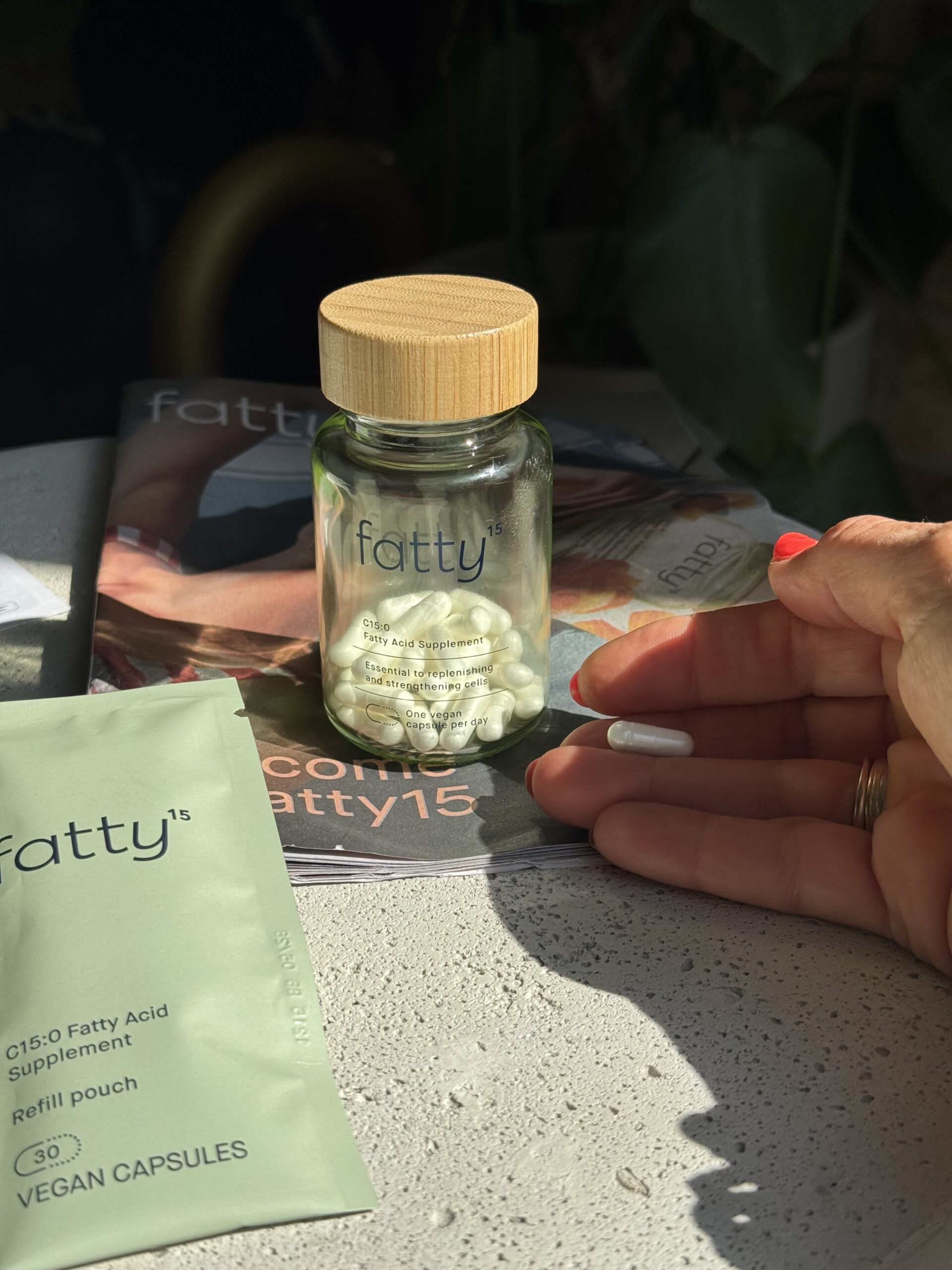 A hand holds a capsule next to a glass jar labeled "fatty15" containing white capsules, with a refill pouch and magazine in the background.