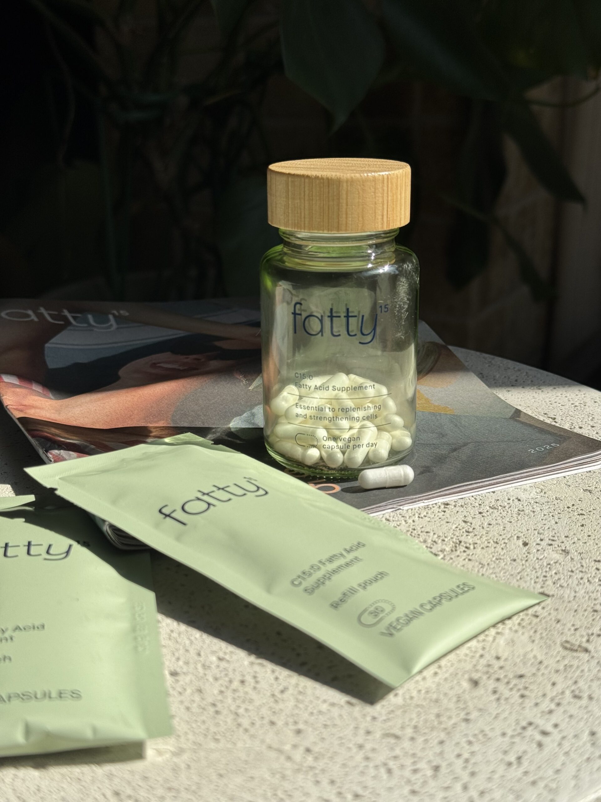 A glass jar labeled "fatty15" filled with white capsules is placed on a magazine, with green supplement packets and a single capsule nearby on a table.