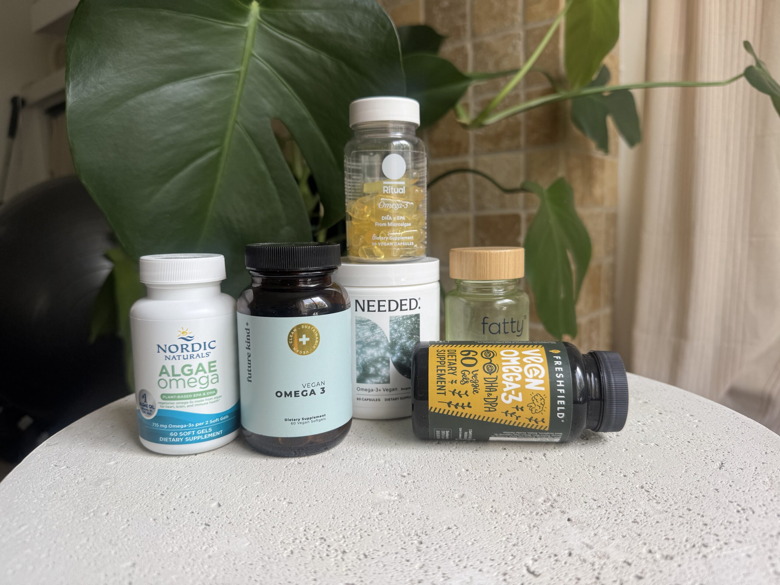 Six omega-3 supplement bottles, including brands like Nordic Naturals and Fatty, are arranged on a white surface with a leafy green plant in the background.