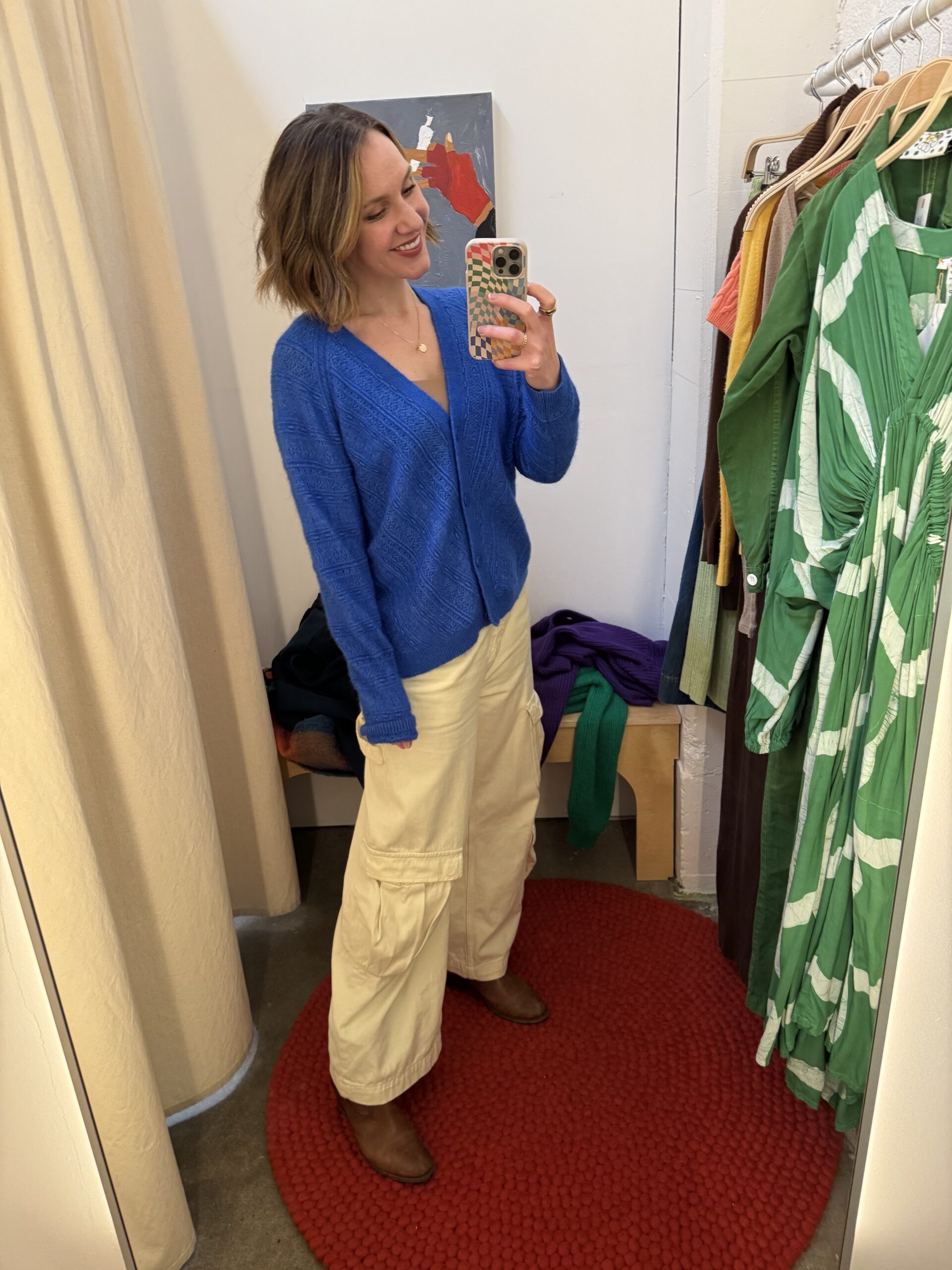 Woman stands on a red circular rug in a fitting room, wearing a blue sweater, cream cargo pants, and brown boots, taking a mirror selfie with her smartphone.
