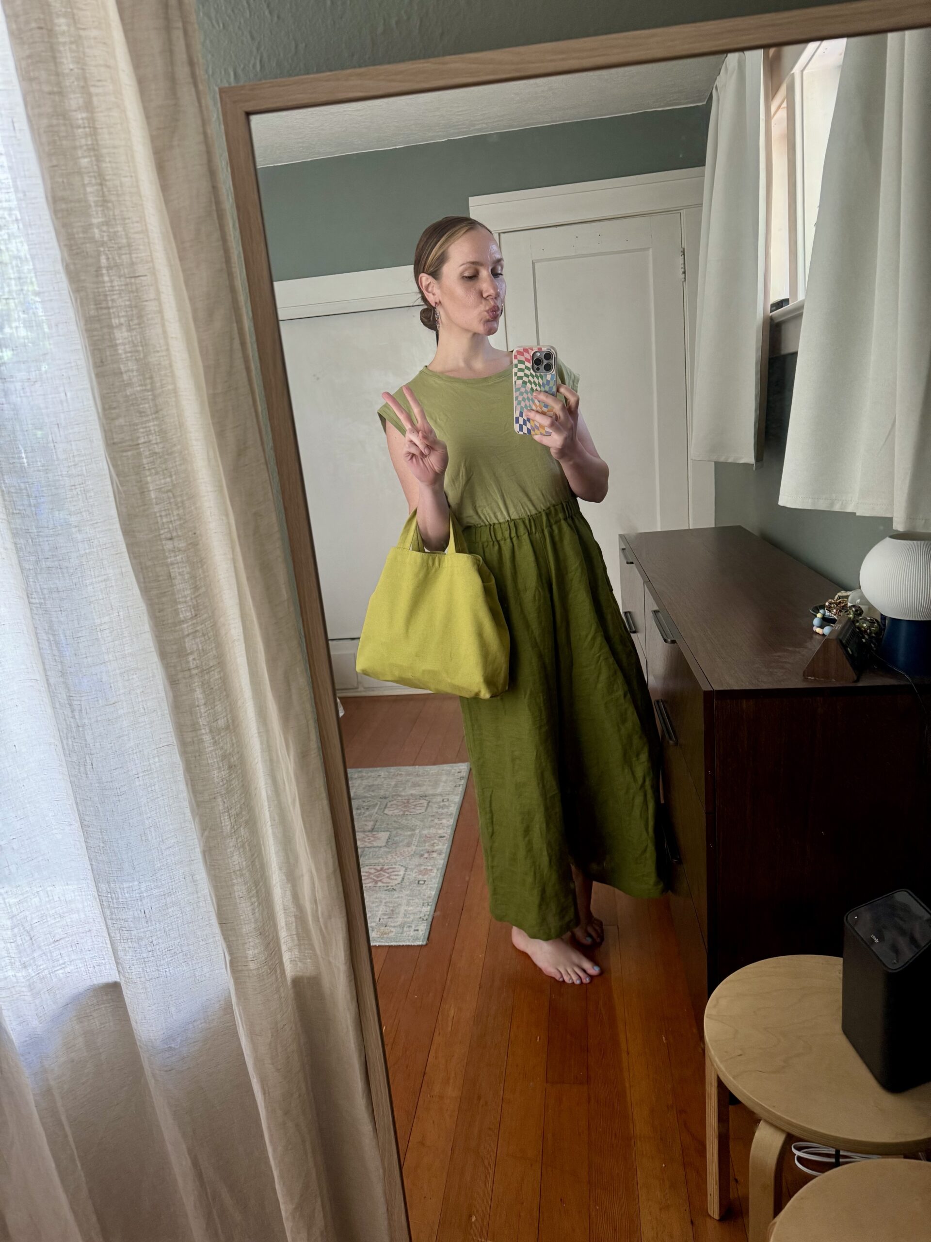 A person stands barefoot in front of a mirror, wearing a green dress and holding a matching green bag, taking a selfie and making a peace sign.