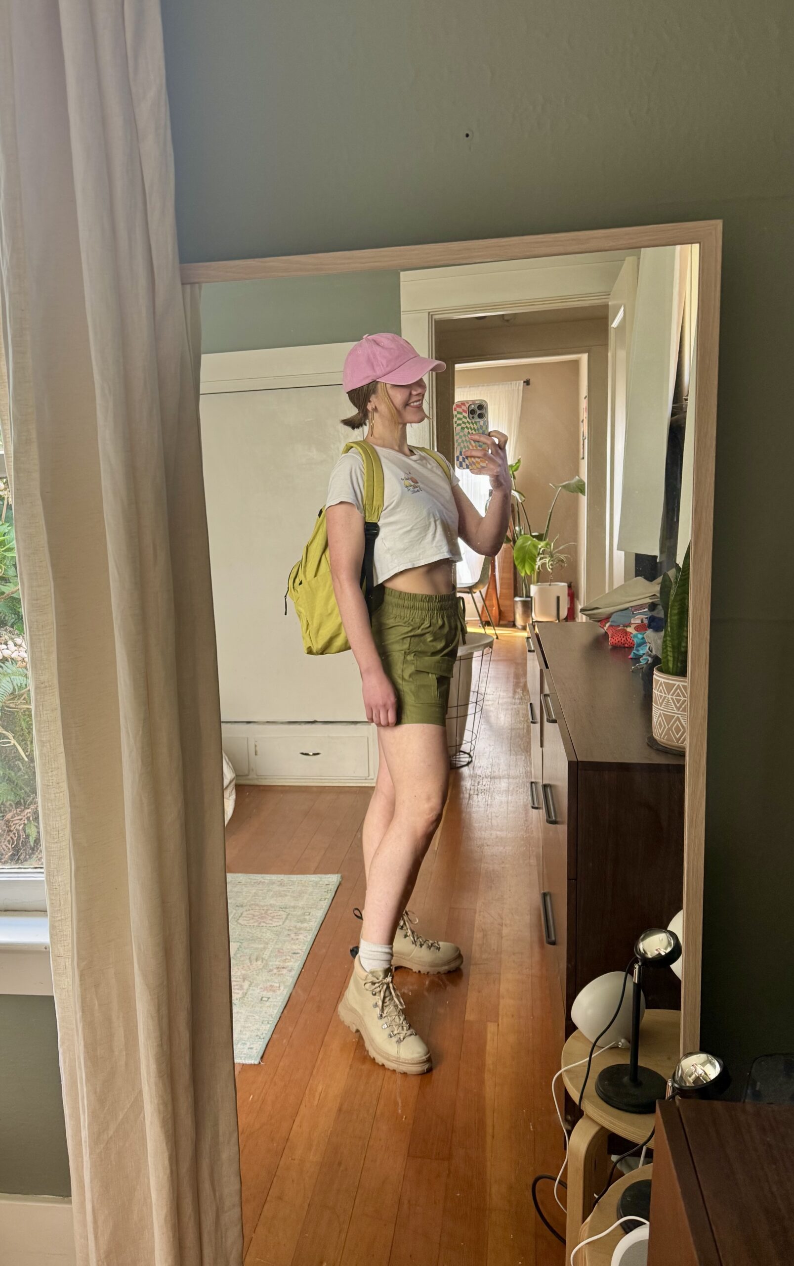 Person taking a mirror selfie in a room, wearing a pink cap, white crop top, green shorts, yellow backpack, and tan boots. The room has wooden floors and furniture.