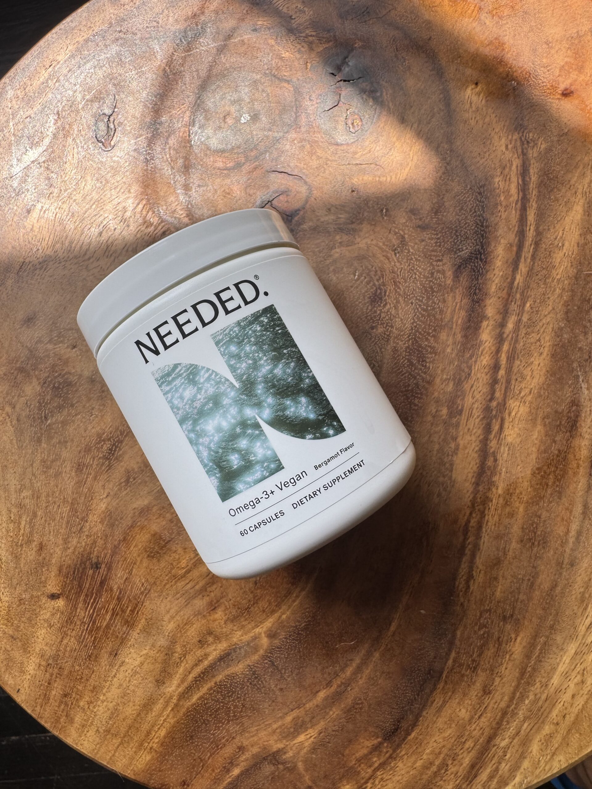 A white container of Needed Omega-3 Vegan dietary supplement capsules rests on a round wooden surface.