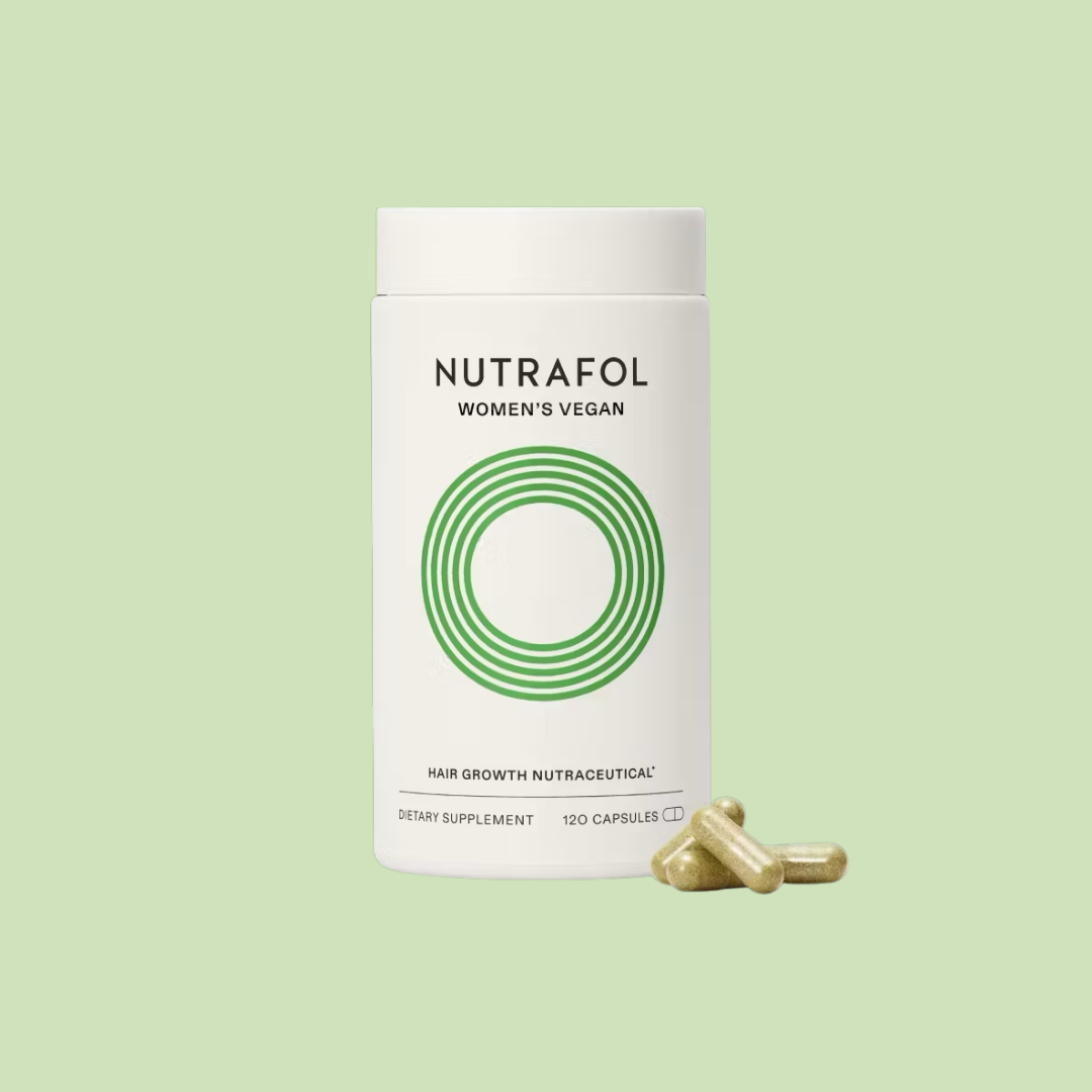 A white bottle labeled "Nutrafol Women's Vegan" hair growth supplement stands upright with three capsules beside it on a light green background.