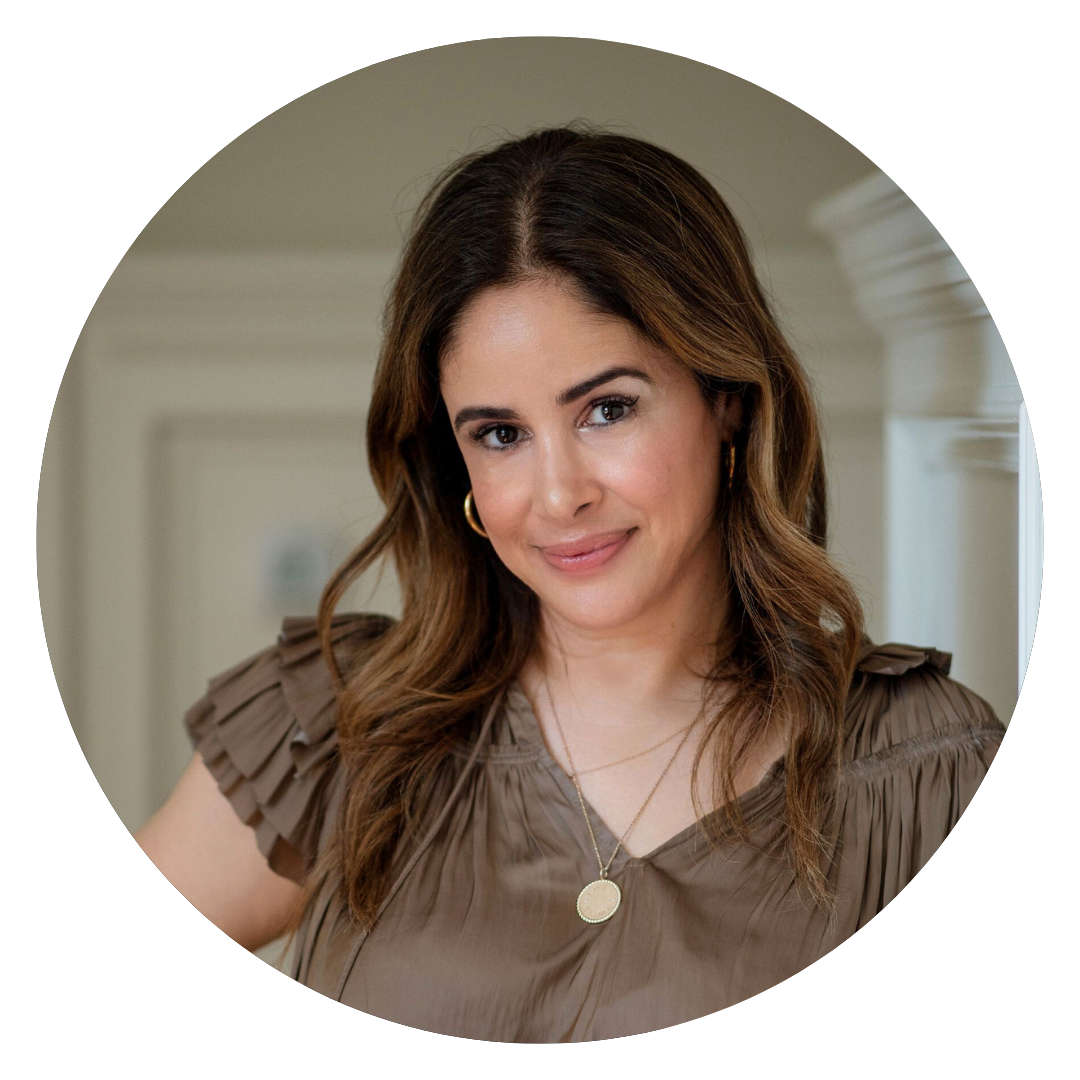 A woman with wavy brown hair, wearing a taupe blouse and gold necklaces, stands indoors and smiles slightly at the camera.
