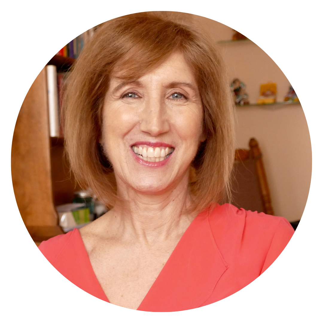 A woman with short, light brown hair and a coral top smiles at the camera in a warmly lit indoor setting.