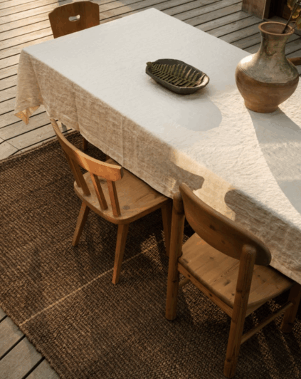 Wooden dining table with a white tablecloth, ceramic vase, and leaf-shaped dish, surrounded by wooden chairs on a textured brown rug.