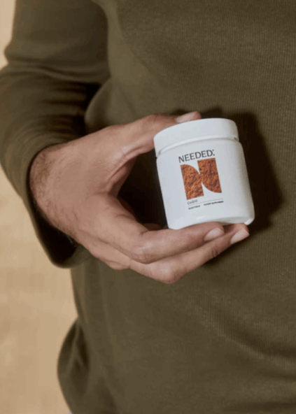 A person wearing an olive green long-sleeve shirt holds a white container labeled "Needed:" with an orange and brown abstract design.