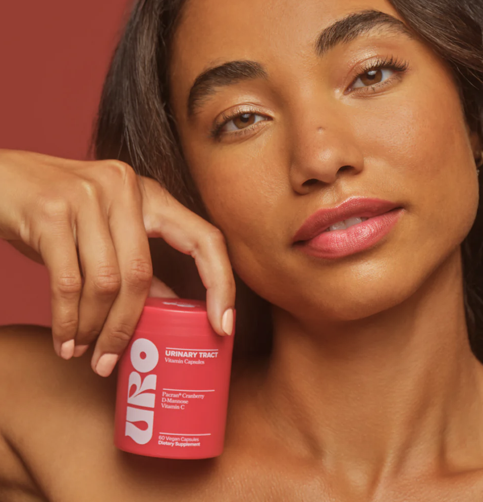 A woman holds a pink container of urinary tract vitamin capsules labeled "URO" close to her face against a solid reddish background.