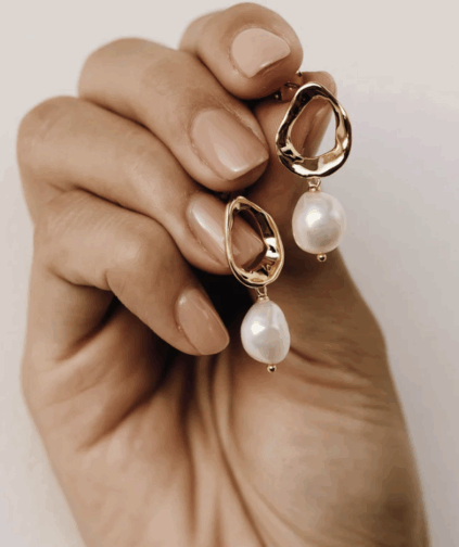 A hand with manicured nails holds a pair of gold earrings featuring irregular open shapes and dangling white pearls.