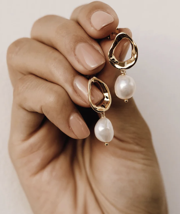 A hand with manicured nails holds a pair of gold earrings featuring irregular open shapes and dangling white pearls.