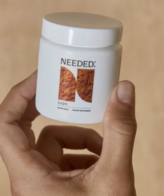 A hand holds a white jar labeled "NEEDED: CoQ10" with a large stylized letter N and text indicating it contains 30 capsules of a dietary supplement.