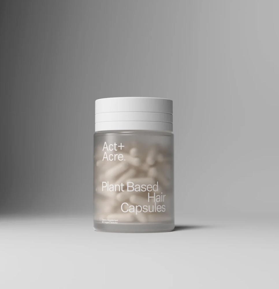 A frosted jar labeled "Act+Acre Plant Based Hair Capsules" is filled with capsules, set against a plain gray background.