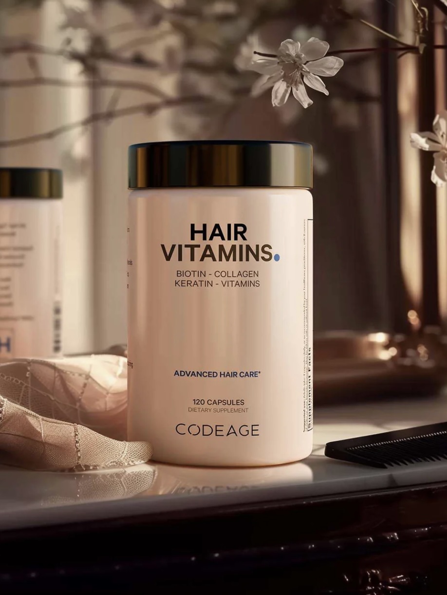 A bottle of Codeage Hair Vitamins, labeled with biotin, collagen, keratin, and vitamins, sits on a counter next to a comb and a flower decoration.
