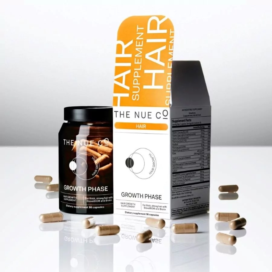 A bottle and box of The Nue Co. Growth Phase Hair Supplement are shown with several capsules scattered around them on a reflective surface.