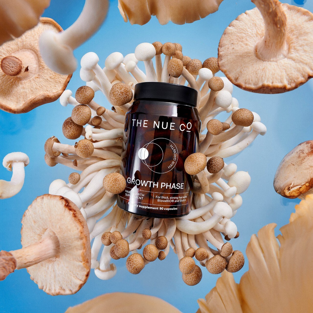 A bottle of The Nue Co. Growth Phase supplements is surrounded by various mushrooms against a blue background.