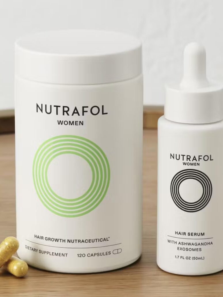 Nutrafol Women hair growth supplement bottle, hair serum dropper bottle, and several yellow capsules on a wooden tray with a hairbrush in the background.