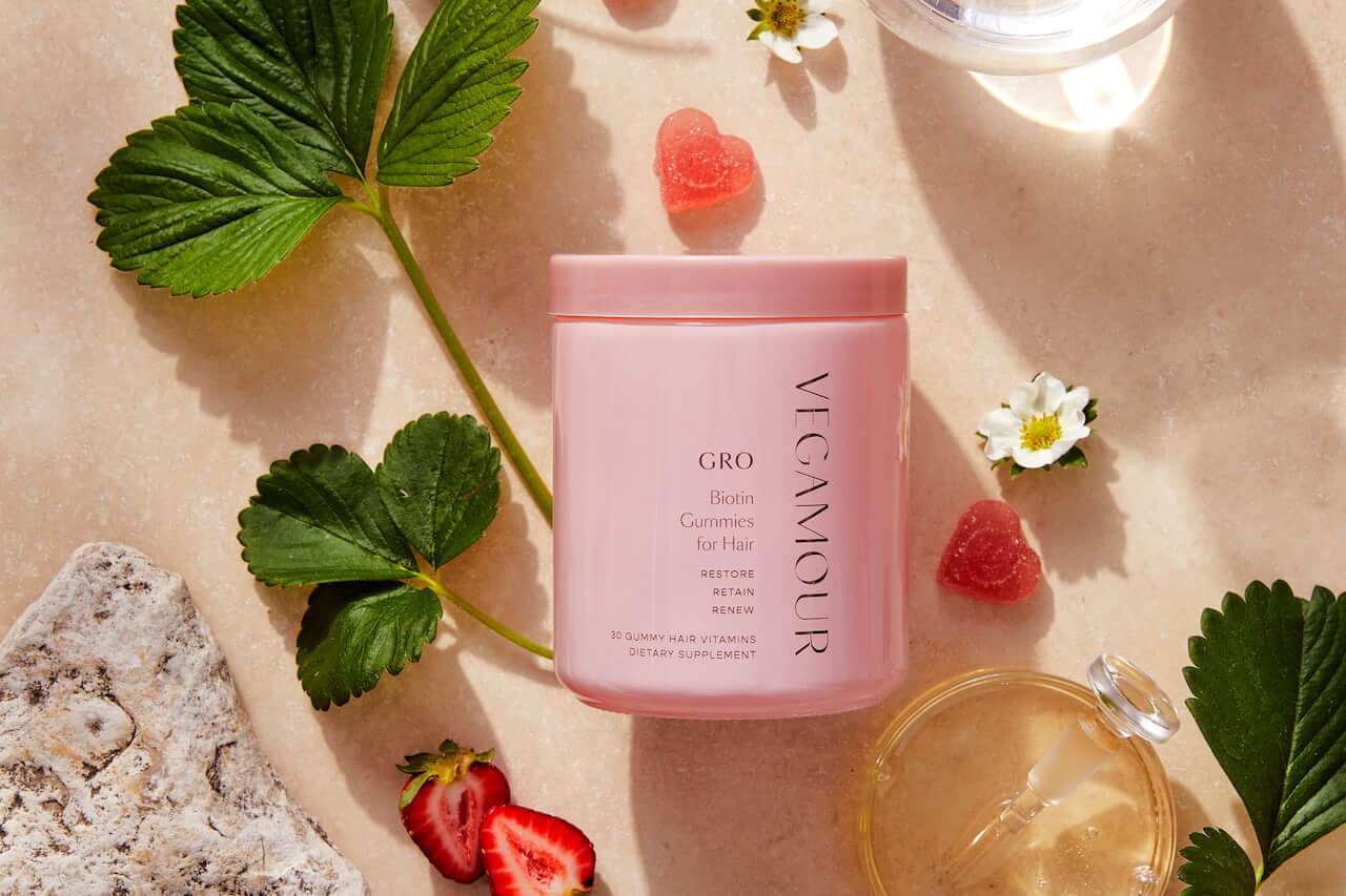 A pink VEGAMOUR GRO Biotin Gummies for Hair container is surrounded by strawberry slices, gummy hearts, green leaves, white flowers, a rock, and a glass cup on a beige surface.